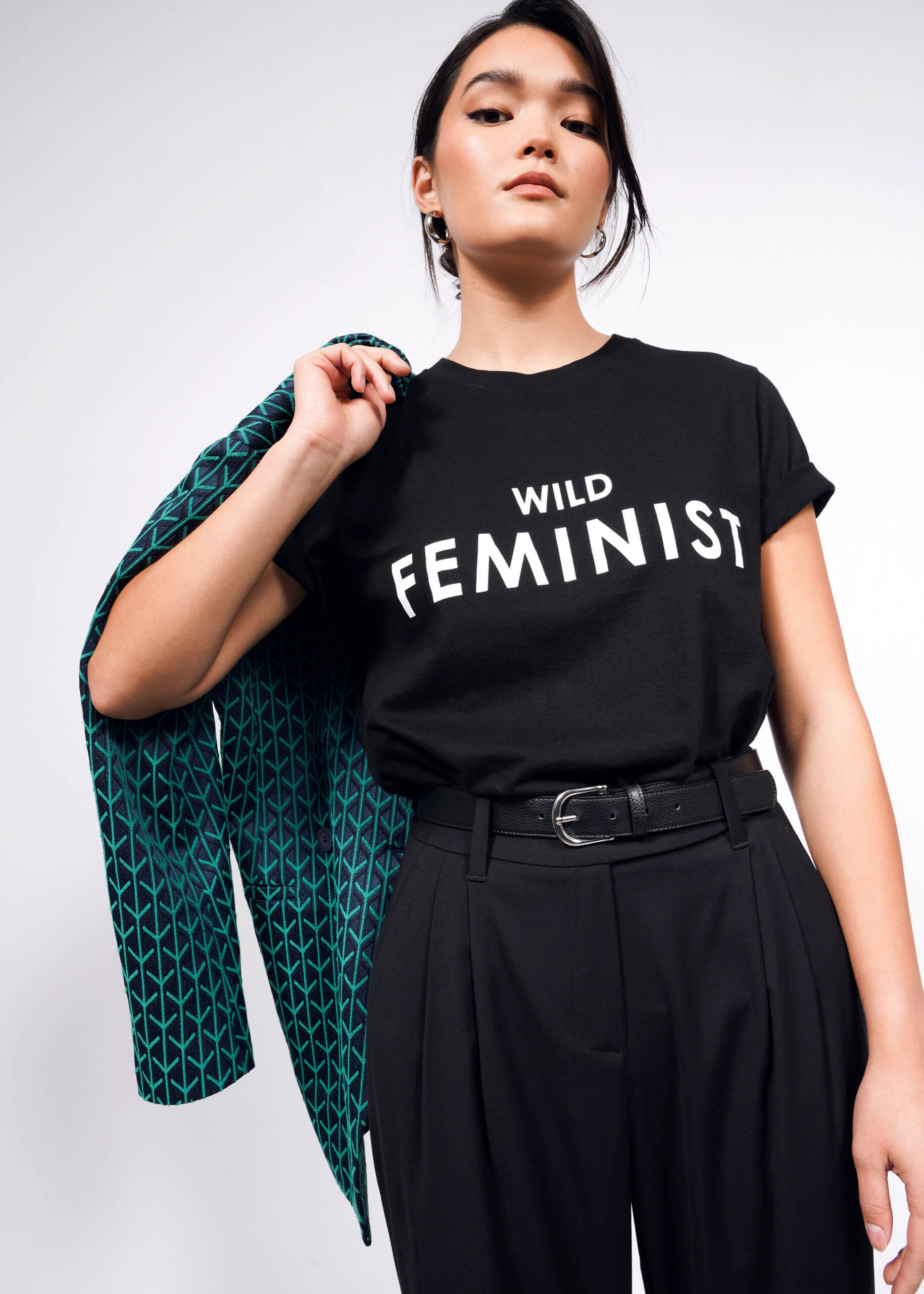 A woman confidently poses in The Wild Feminist® Tee, a black unisex fit T-shirt with WILD FEMINIST in bold white letters, made from pre-shrunk 100% cotton for lasting comfort. She holds a jacket over her shoulder and pairs it with high-waisted black pants against a plain white background.