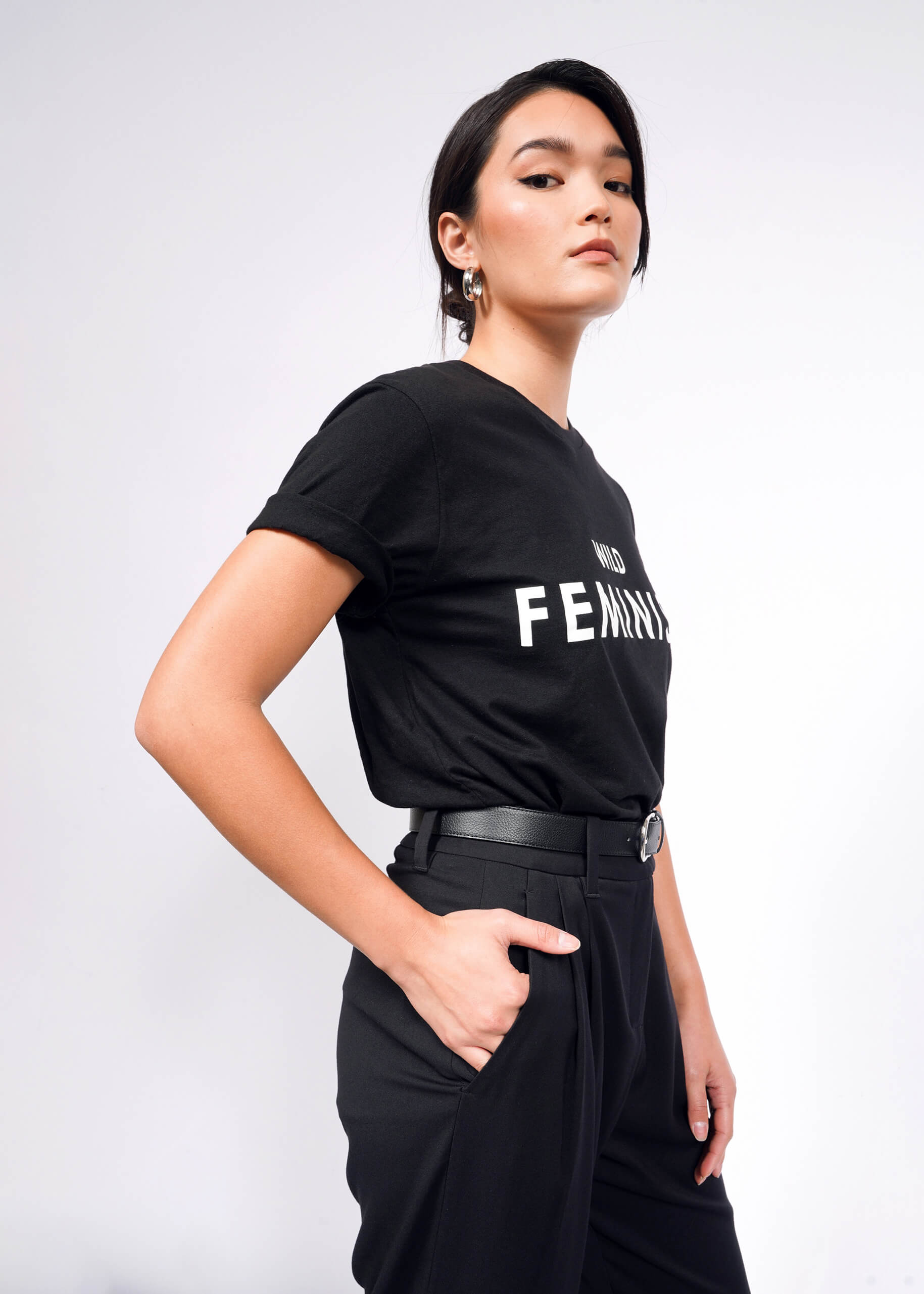 Confidently standing against a plain backdrop, a short-haired woman with hoop earrings wears The Wild Feminist® Tee, a unisex black cotton t-shirt with FEMINIST boldly displayed. Her hand in her pocket complements her bold stance.