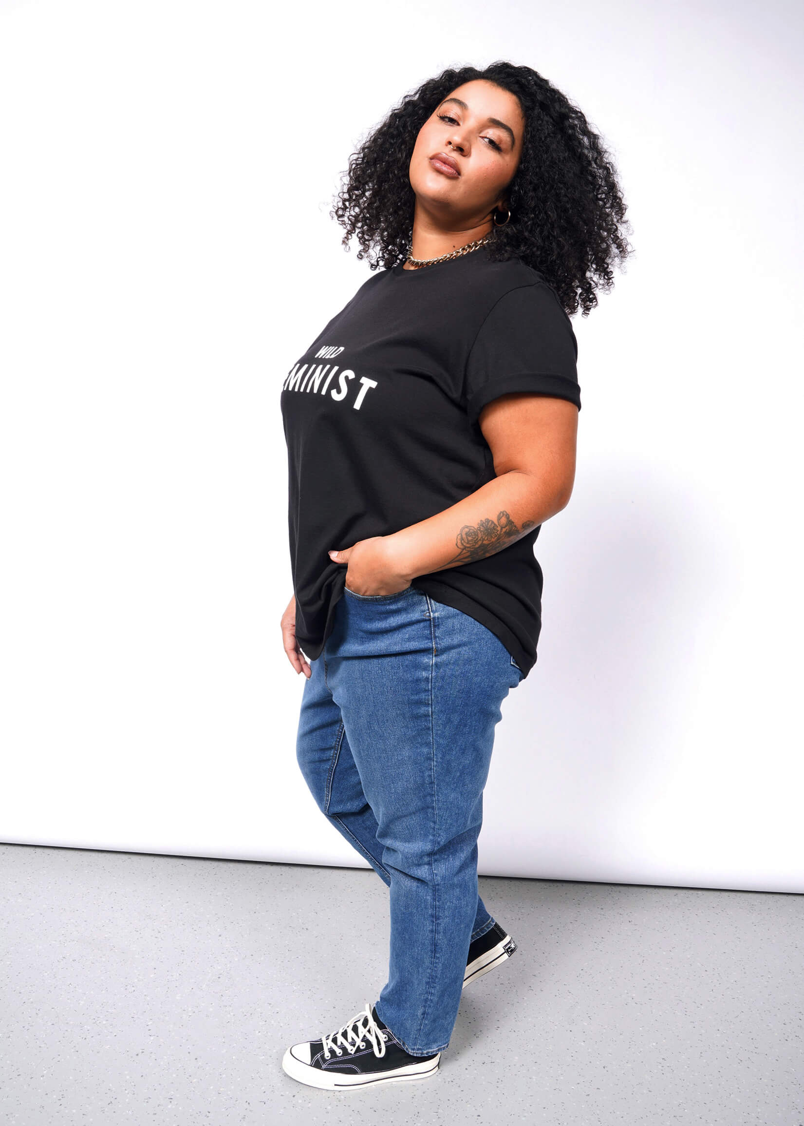 A person with curly hair confidently wears The Wild Feminist® Tee, a black cotton unisex shirt with Feminist printed on it. Paired with blue jeans and black sneakers, they pose boldly against a plain white background.