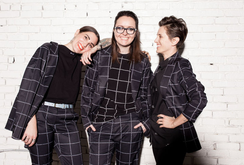 Three women wearing black suits and smiling while standing in front of white brick wall.