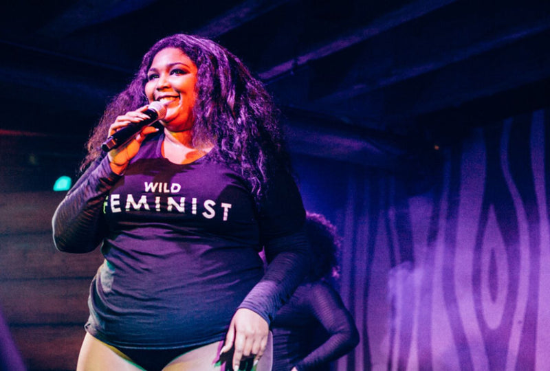 Lizzo wears  black Wild Feminist tee while on stage with microphone