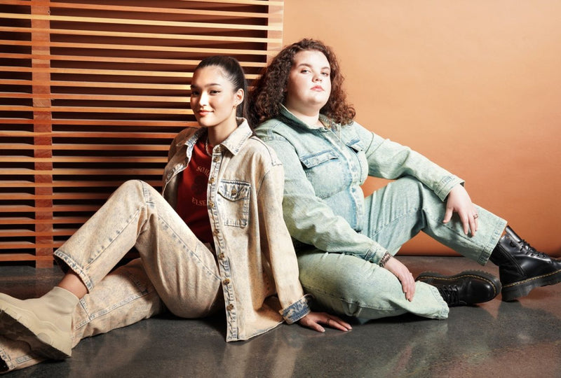 Two people sit back to back in front of orange and wood backdrop wearing denim outfits and boots.