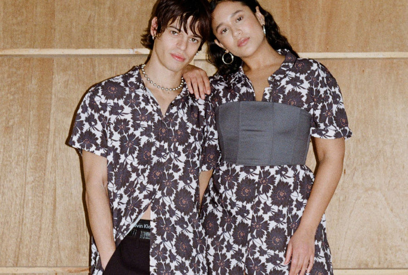 Two models wear floral printed button up dress in front of light wood backdrop.