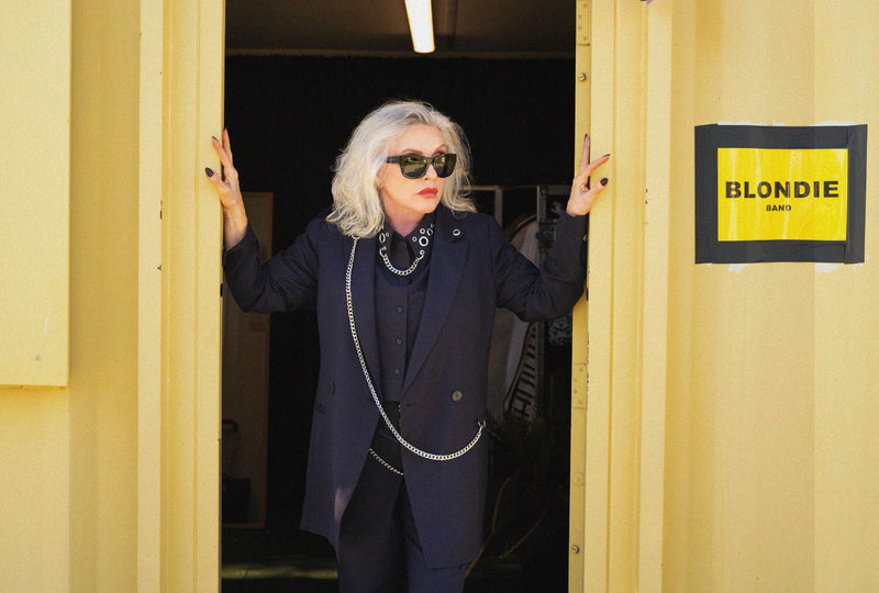 Debbie Harry wears black suit while standing in yellow doorway next to sign that reads "Blondie band"