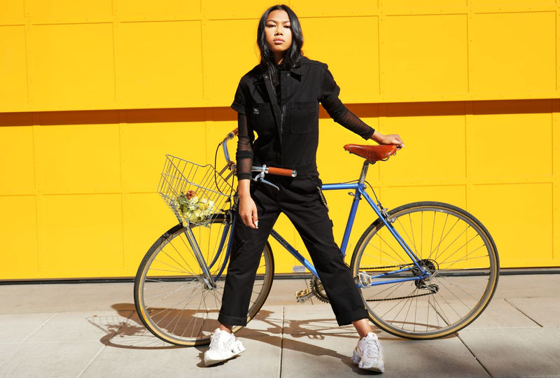 Against bright yellow garage door, person wearing black coverall stands in front of bike while looking at camera.