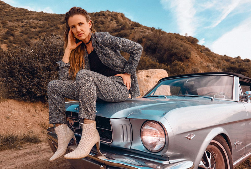 Juliette Lewis wears a teal patterned suits and white boots while sitting on the hood of blue vintage car..
