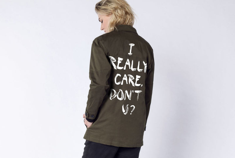 Against white background, person wears green jacket with text on the back that reads "I really care, don't u?"