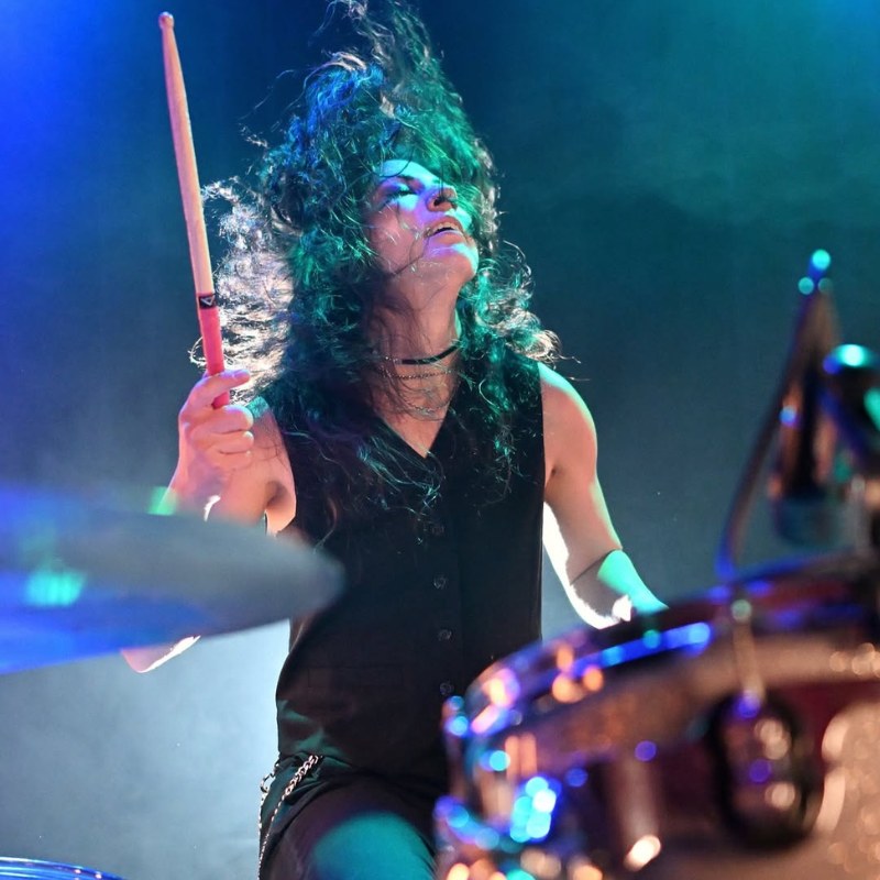 Person playing drums wearing black vest.