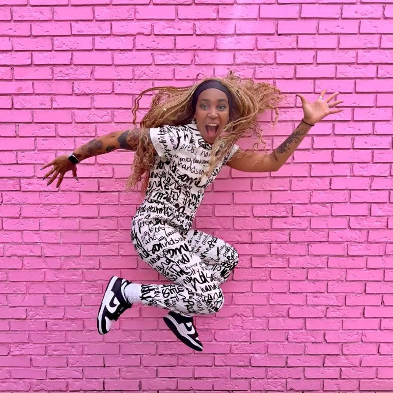 Person wearing black and white jumpsuit while jumping in the air in front of pink brick background.