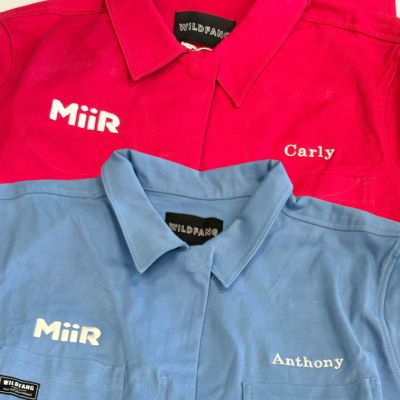 Two coveralls, pink and blue, lay flat on top of each other showing white custom embroidery