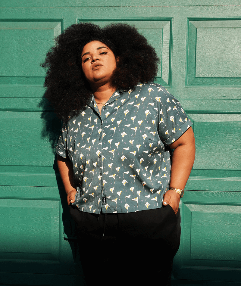 Model with black hair stands in front of green paneled wall with their hands in their pant pockets wearing The Everyday Camp Button Up in Calallilies Dark Dark.