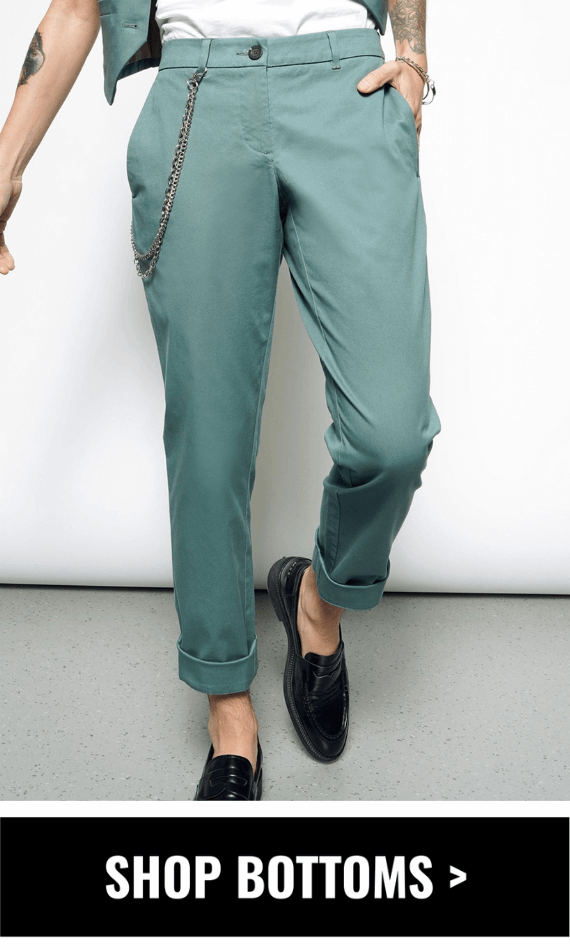 A person stands casually with a tattooed arm, hand in their pocket, wearing The Essential Trouser in dark teal with a chain accessory, paired with a white shirt and cropped teal vest. They wear black loafers against a plain white background. Underneath image, black button reads SHOP BOTTOMS