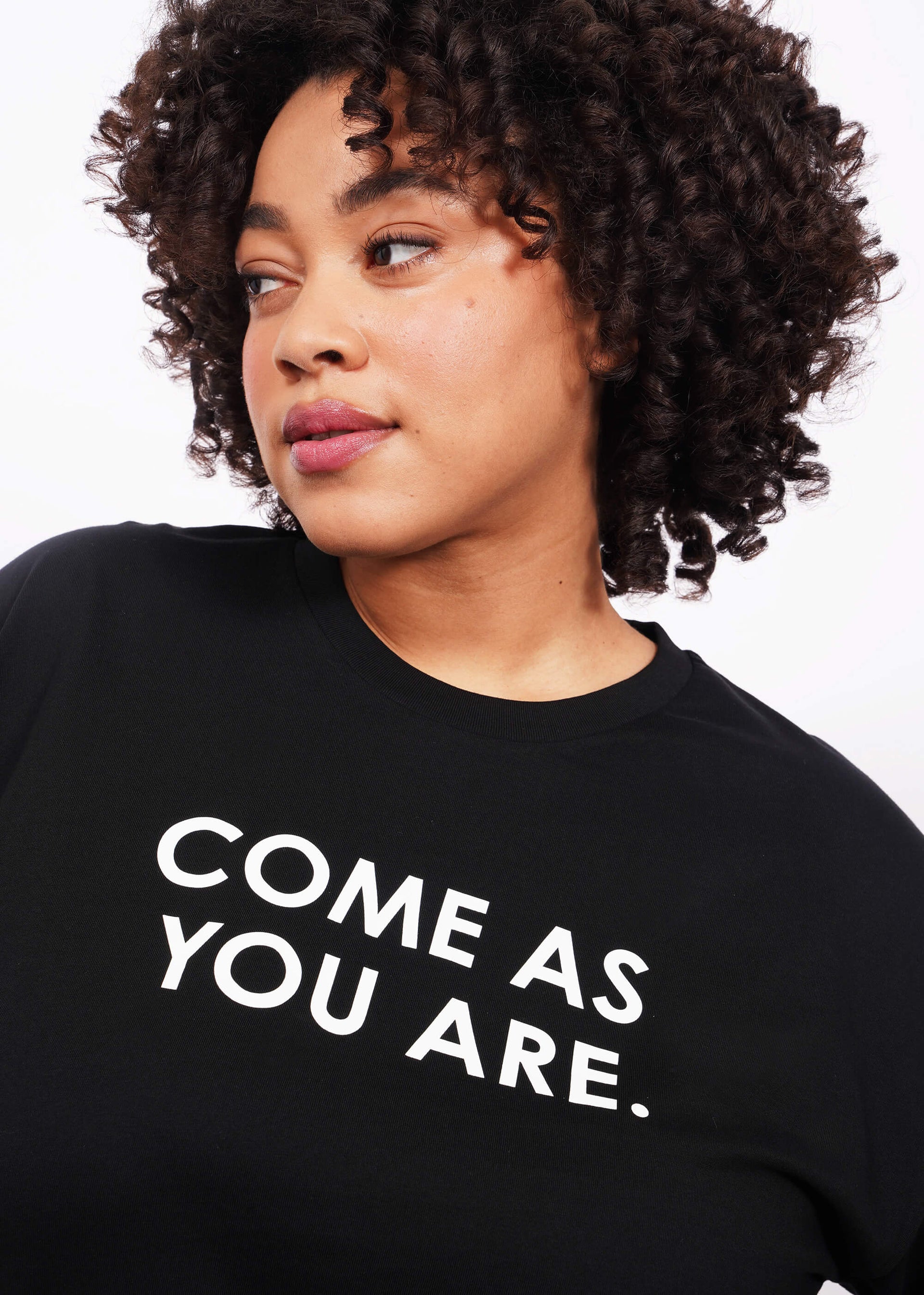 Close up, chest up view of model with short curly hair wearing The Come As You Are Tee in black, which is a black relaxed fit tshirt with bold text across the chest that says "COME AS YOU ARE."