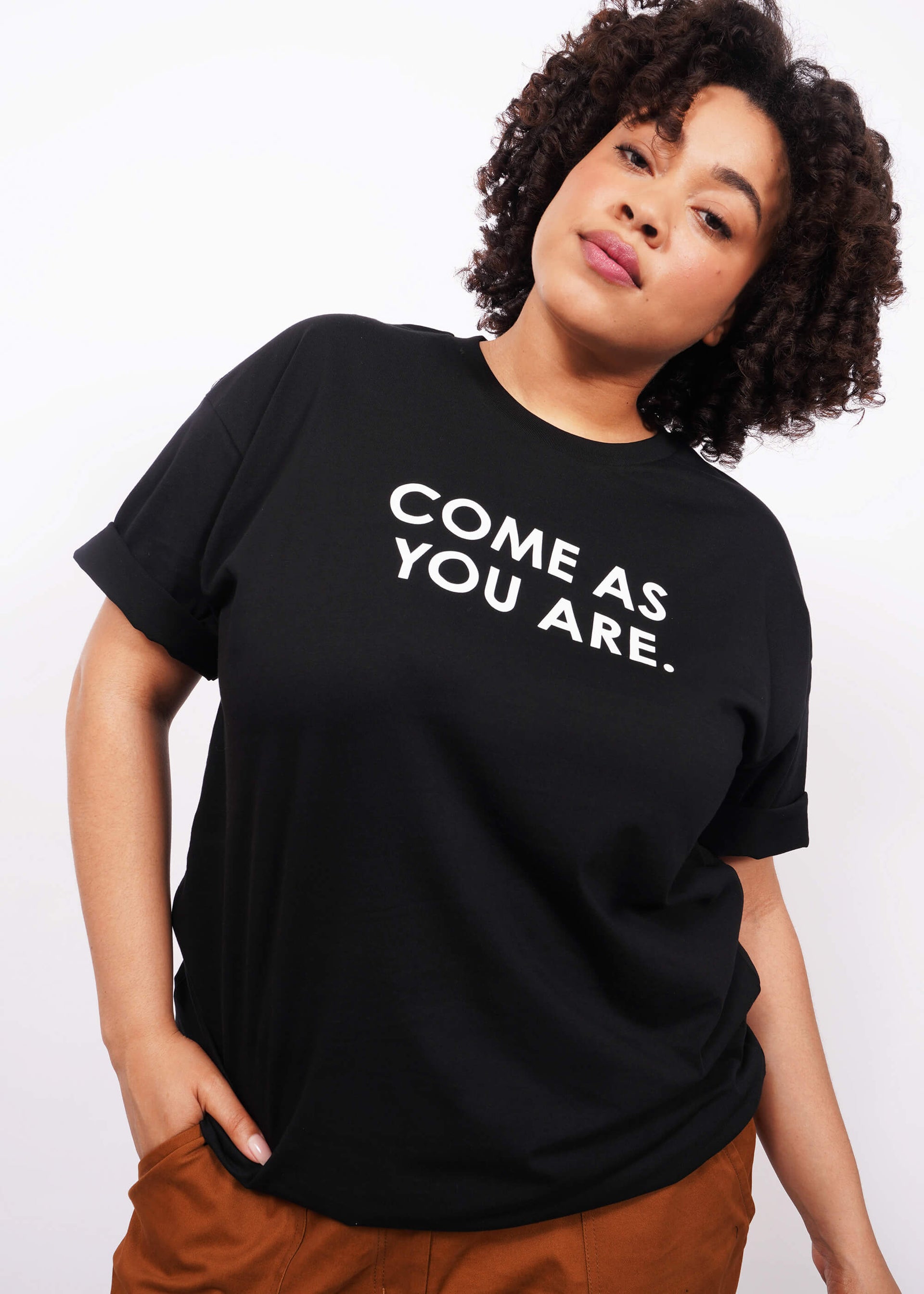 Waist up image of model with short curly hair facing camera wearing The Come As You Are Tee in black, which is a black relaxed fit tshirt with bold text across the chest that says "COME AS YOU ARE."
