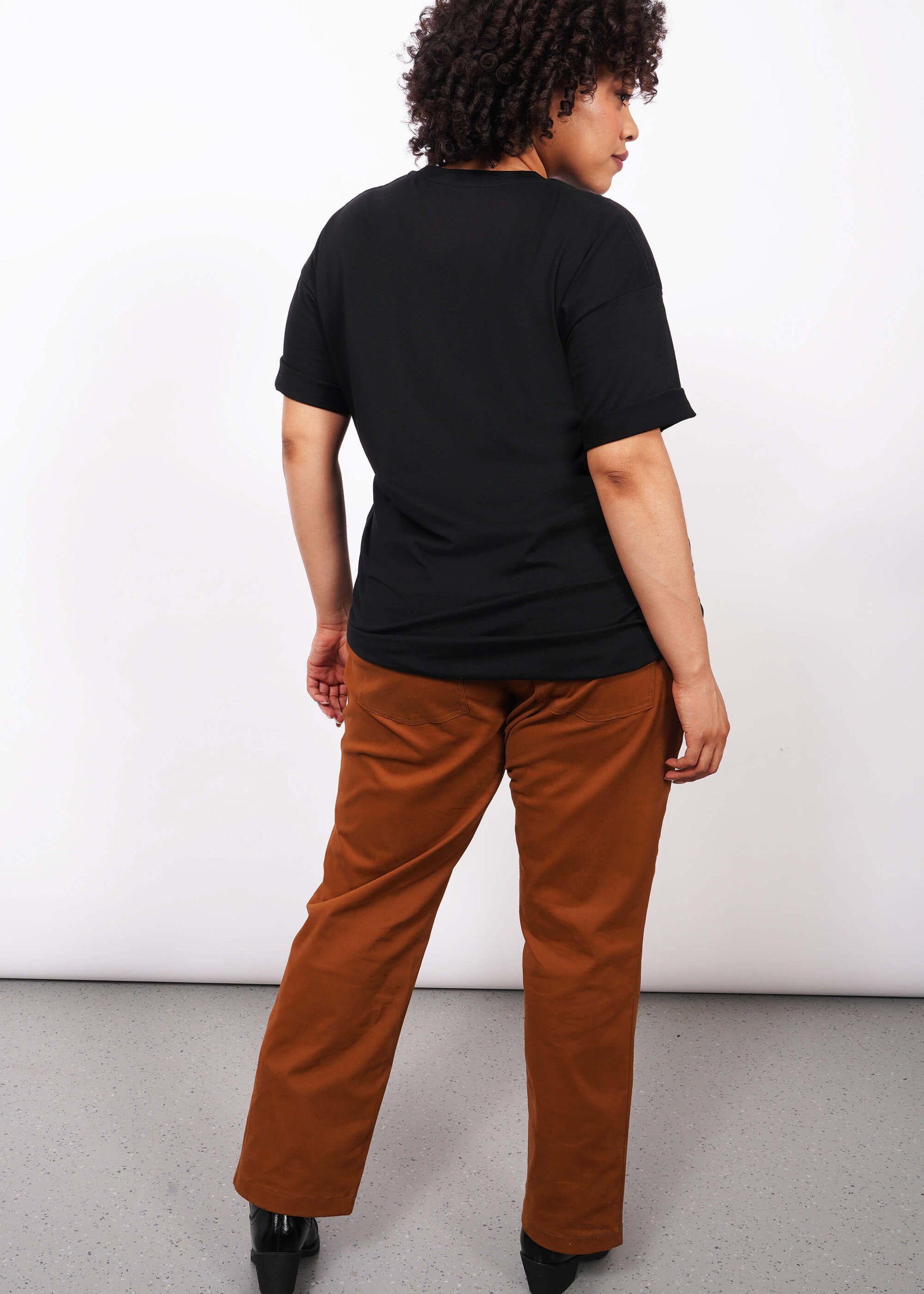 Full body back view image of model with short curly hair wearing brown pants and The Come As You Are Tee in black, which is a black relaxed fit tshirt with bold text across the chest that says "COME AS YOU ARE." The text is not visible in this image since this is a back view of the shirt.