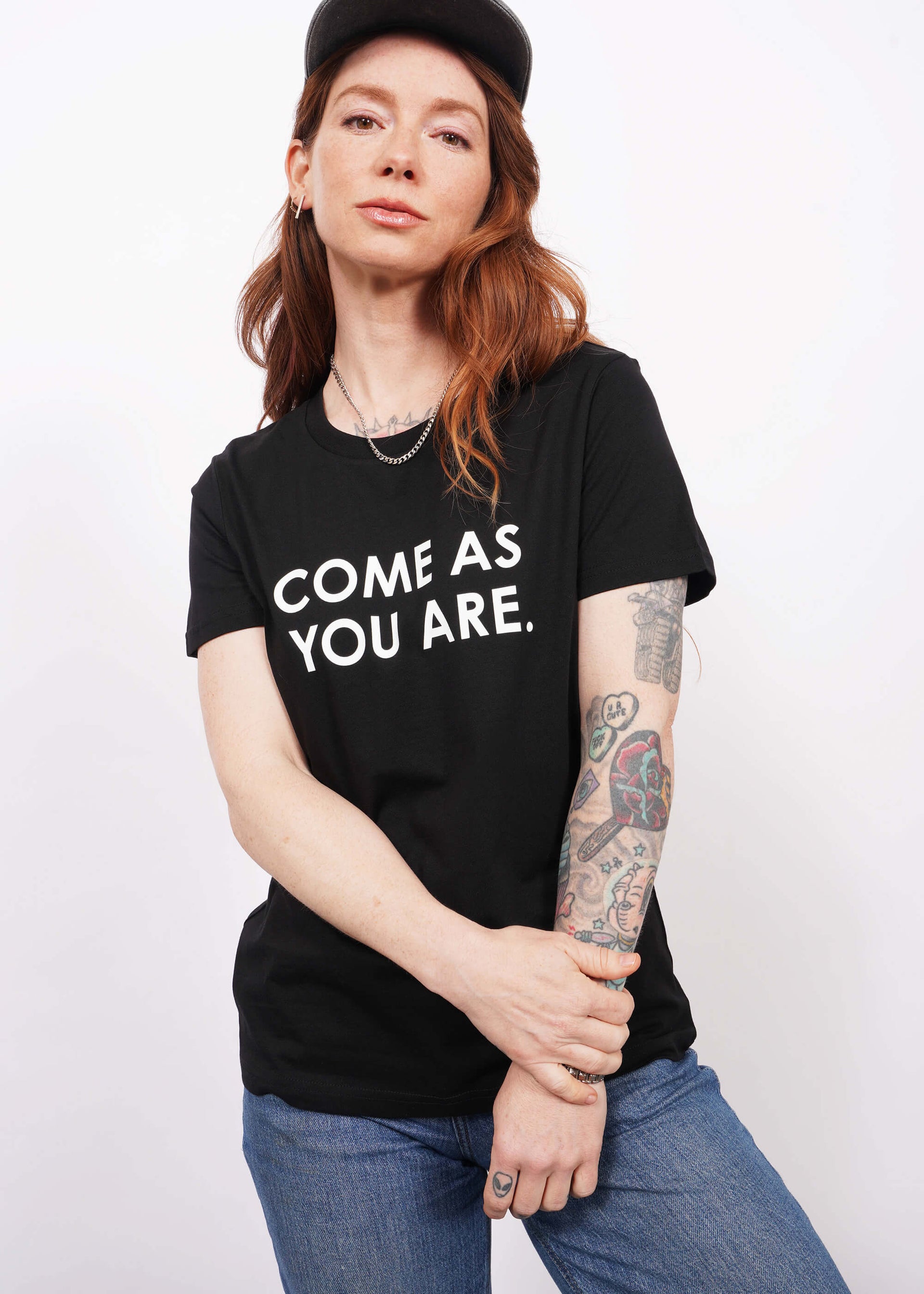 Model with wavy long red hair faces camera wearing a black hat and The Come As You Are Tee in Black, which is a black relaxed fit tshirt with bold white text across the chest that says "COME AS YOU ARE."