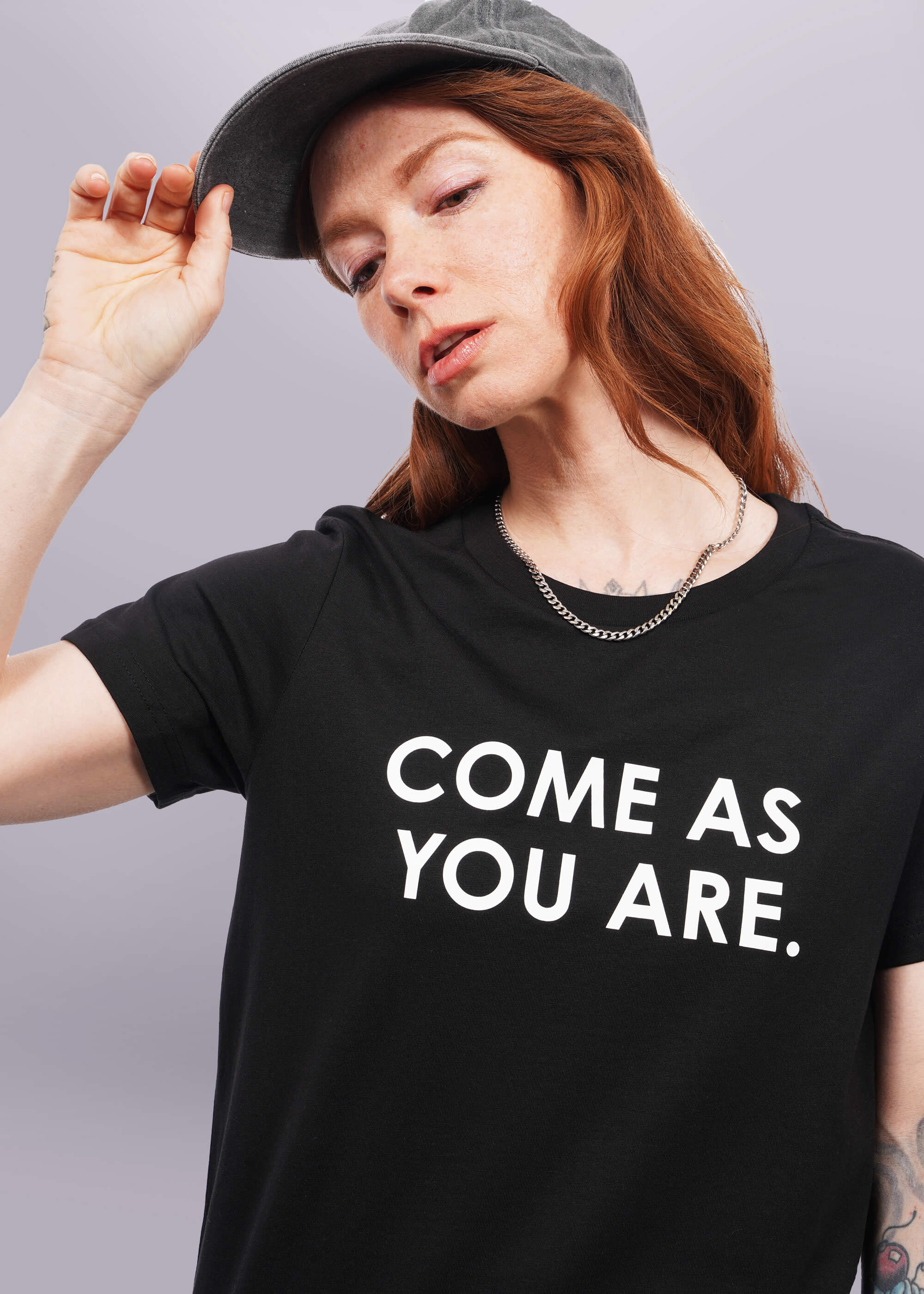 Close up of model with wavy long red hair facing camera wearing a cap and The Come As You Are Tee in Black, which is a black relaxed fit tshirt with bold white text across the chest that says 