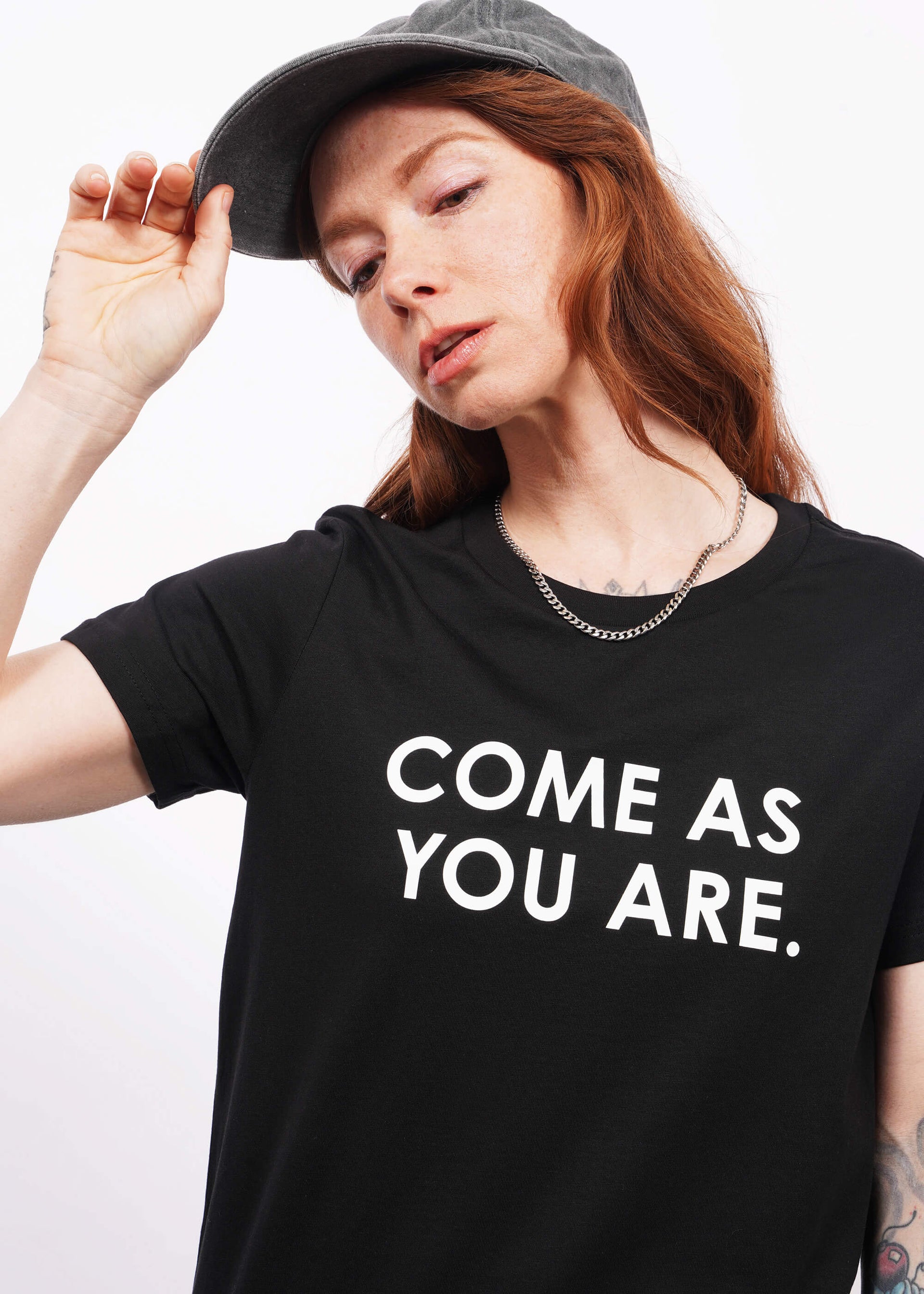 Close up of model with wavy long red hair facing camera wearing a cap and The Come As You Are Tee in Black, which is a black relaxed fit tshirt with bold white text across the chest that says "COME AS YOU ARE."