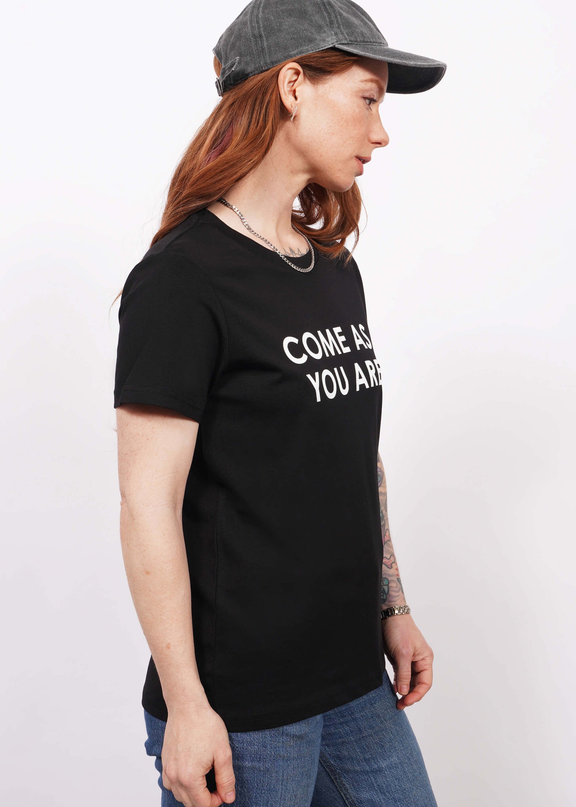 Waist up side view of model with wavy long red hair wearing a cap, jeans, and The Come As You Are Tee in Black, which is a black relaxed fit tshirt with bold white text across the chest that says "COME AS YOU ARE."