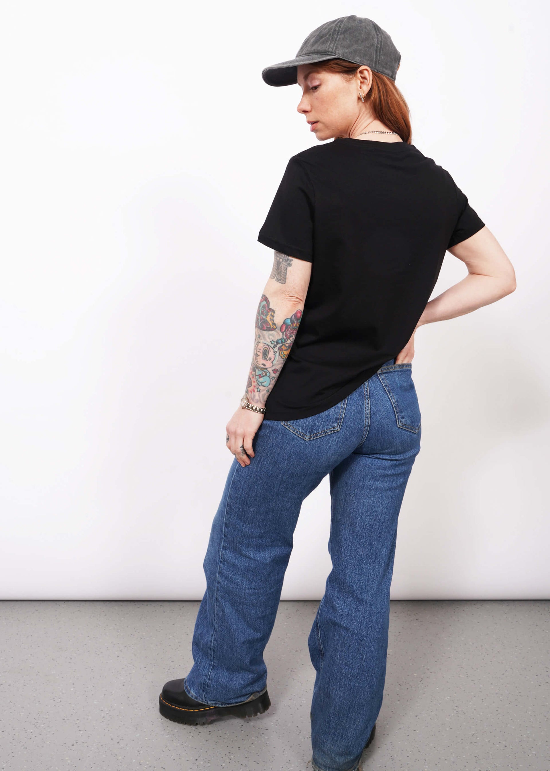 Full body back view of model with wavy long red hair wearing a cap, jeans, and The Come As You Are Tee in Black, which is a black relaxed fit tshirt with bold white text across the chest that says "COME AS YOU ARE." Text is not visible in this view since it shows the back of the shirt.