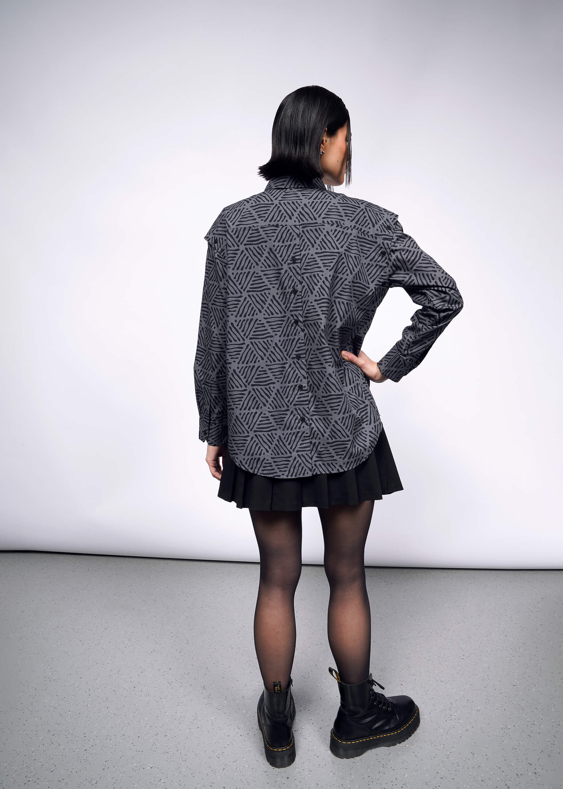 A person with short black hair faces away, wearing The Essential Convertible Sleeve Button Up in gray, a black skirt, sheer tights, and black boots with yellow stitching. The background is a simple light-colored studio setting.