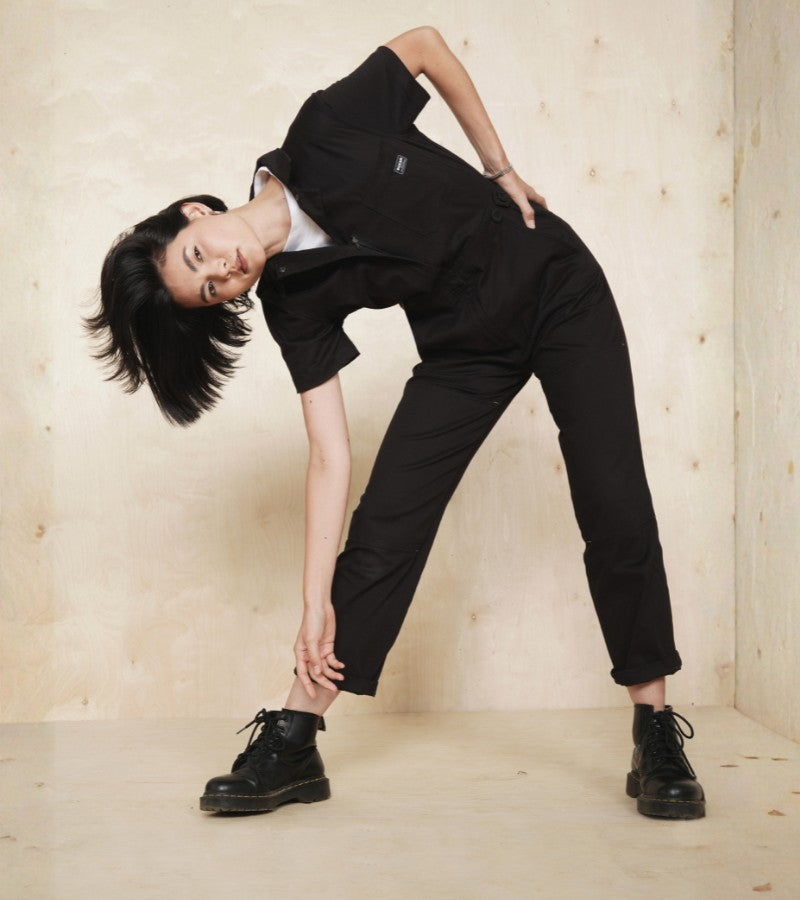 Against light wood backdrop, model wears black coverall and boots leaning over touching ankle.