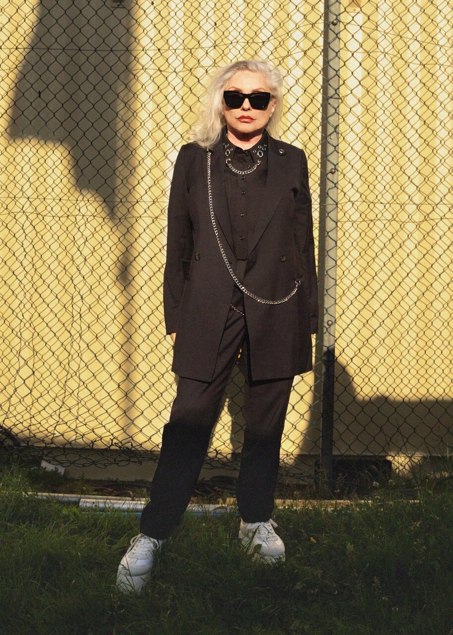 A person with long white hair stands confidently before a chain-link fence, embodying the punk aesthetic. Dressed in Debbie Harry X Wildfang Slim Crop Pants, they accessorize with chains, black sunglasses, and white sneakers. A sunlit yellow wall behind them casts intriguing shadows.