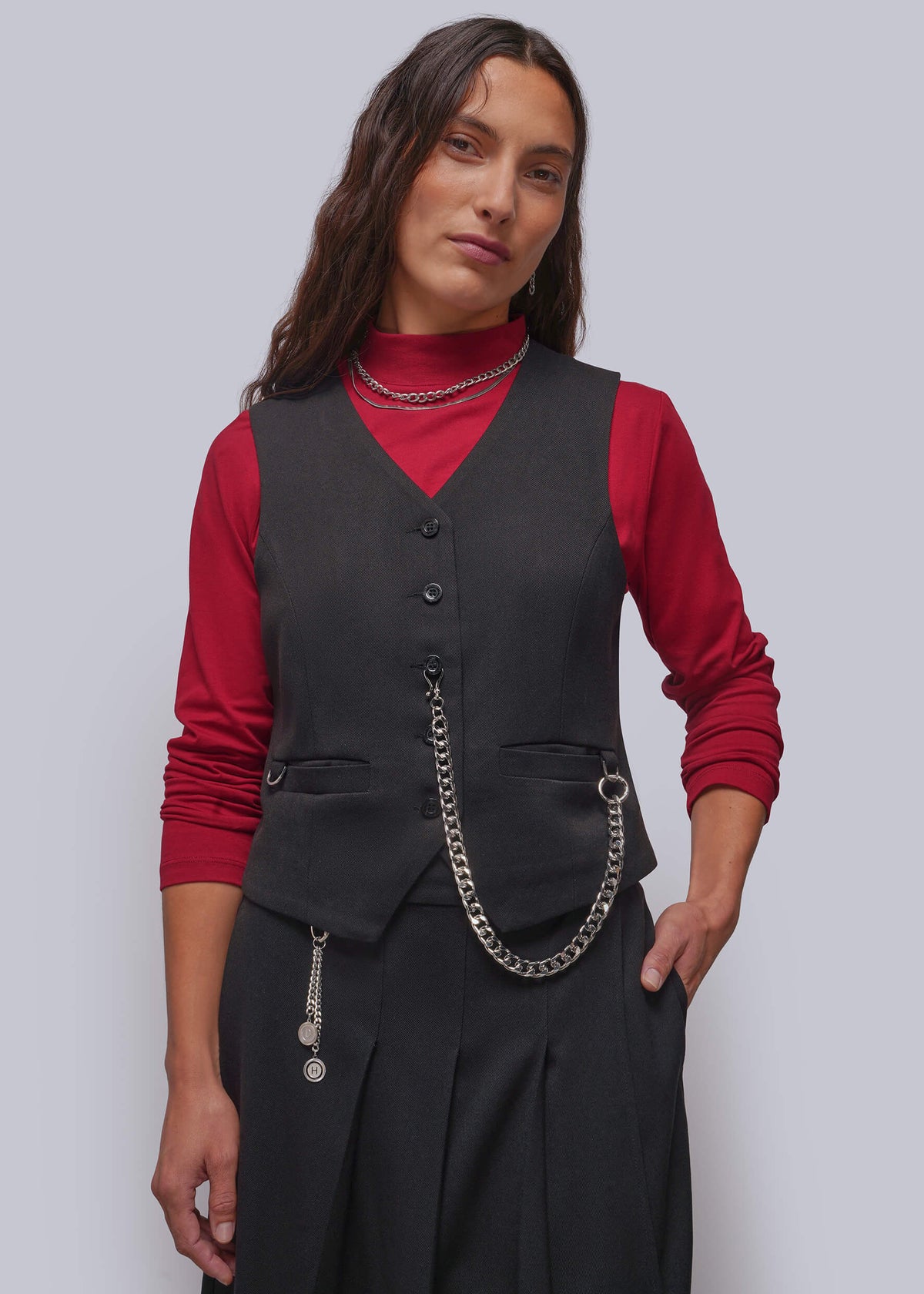 S=A woman modeling a black vest over a red long-sleeve top and black pleated skirt, accessorized with a silver chain belt and layered necklaces, against a solid gray studio background.