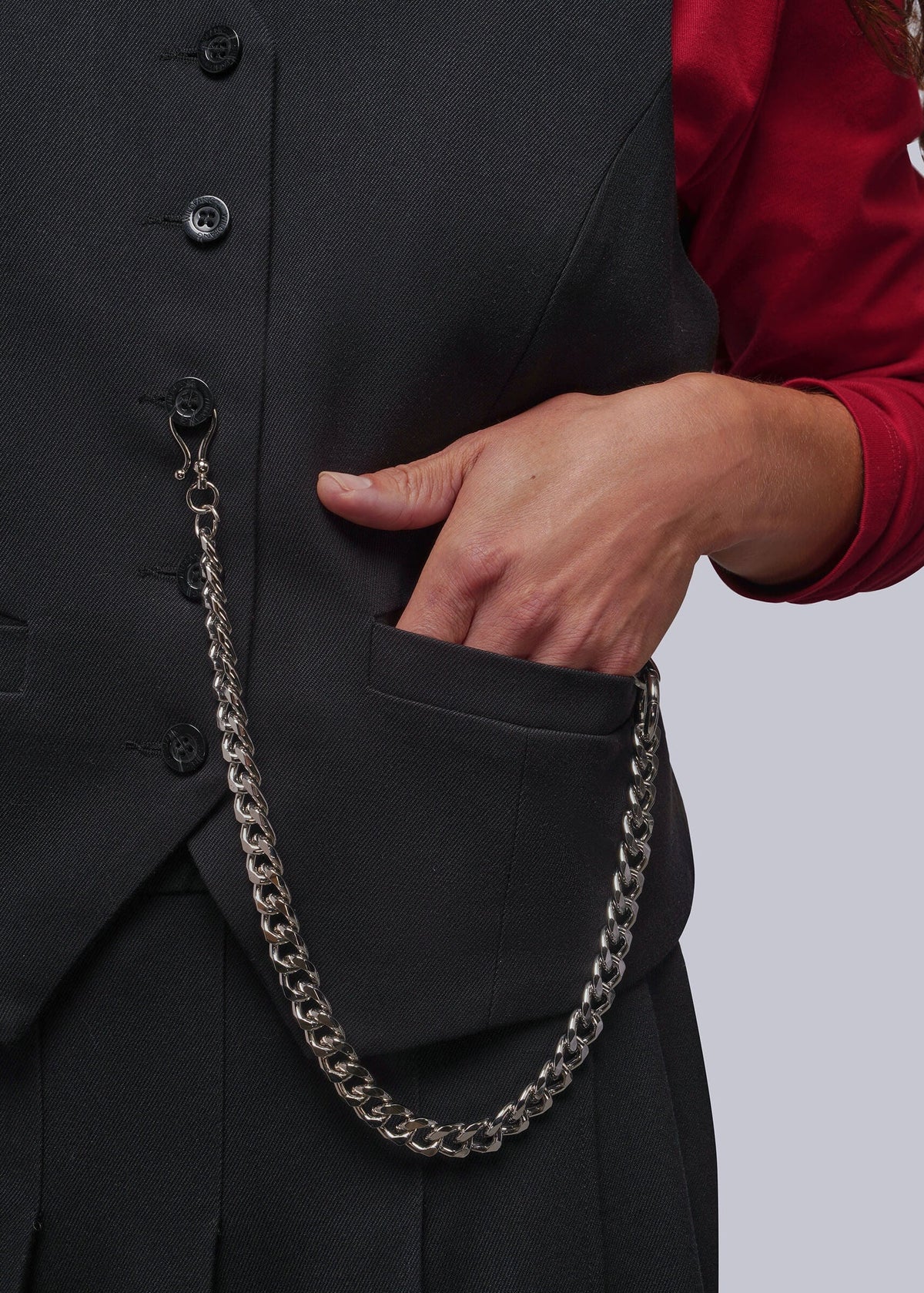 S=A close-up of a person wearing a black vest and skirt, with a silver chain accessory draped from the vest pocket over a red long-sleeve top, against a light gray background.