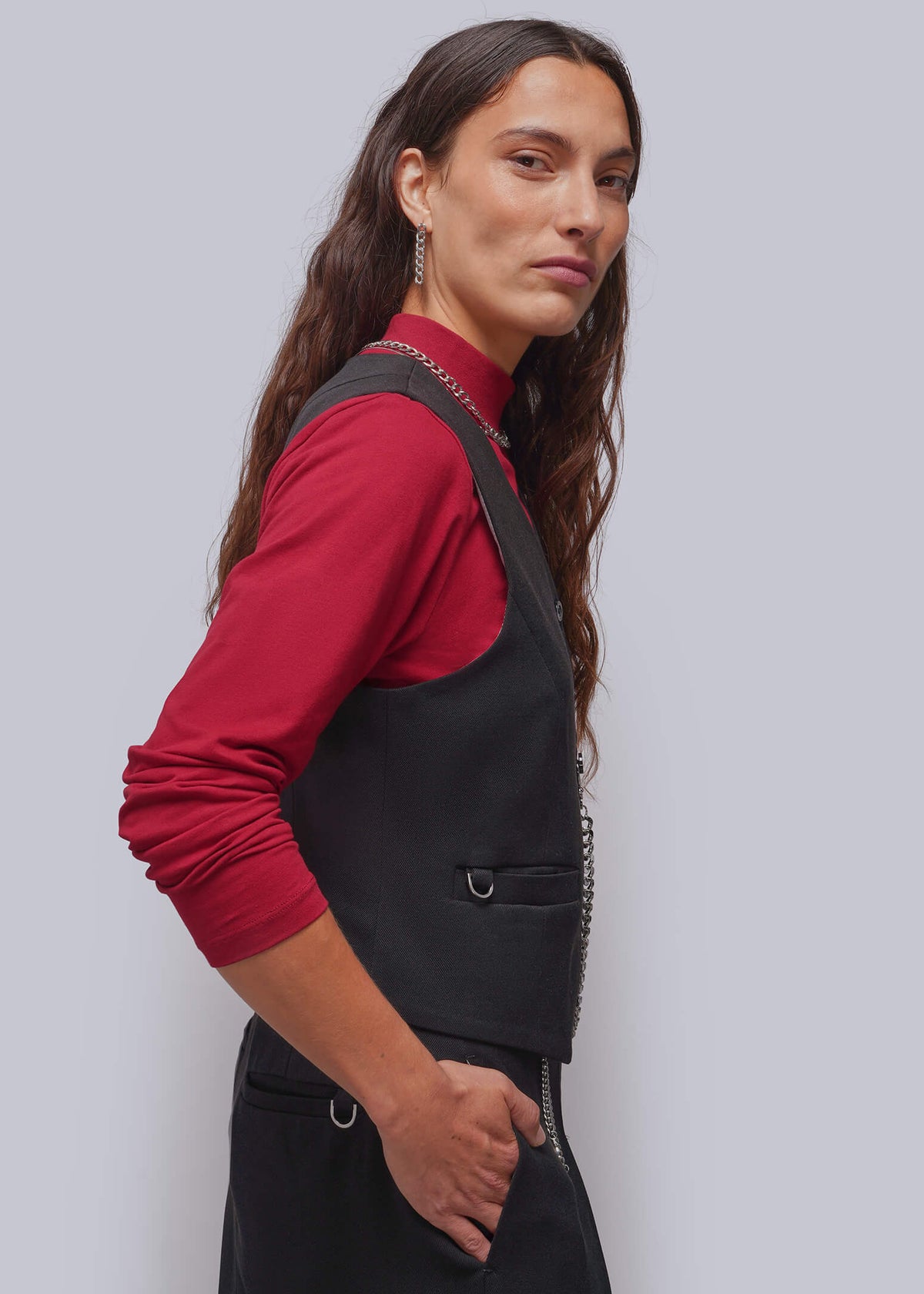 S=A woman modeling a black sleeveless vest over a red long-sleeve mock neck top, photographed against a solid light gray background.