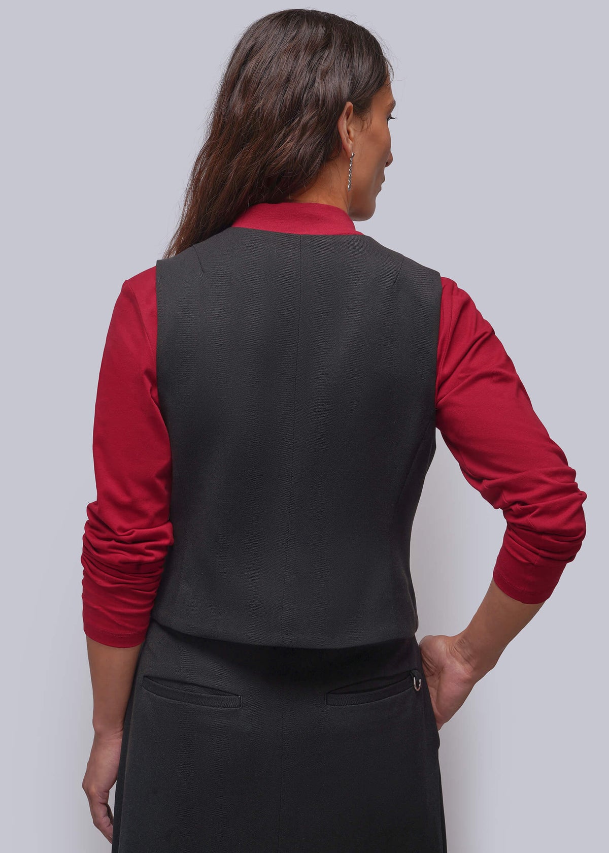 S=A woman shown from the back wearing a black vest over a red long-sleeve shirt and a black skirt, against a plain light gray studio background.