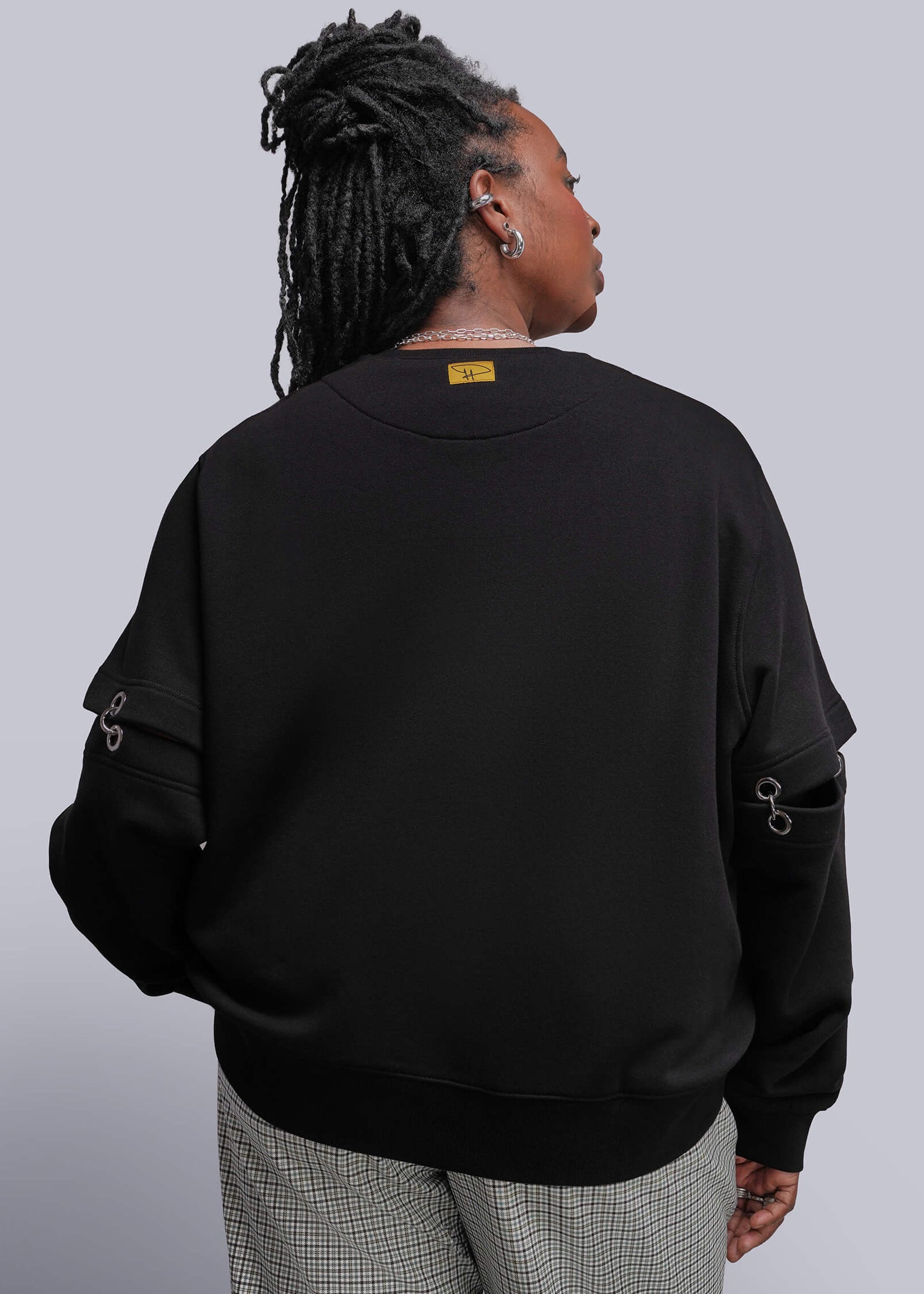 1X=A person shown from the back wearing a black crewneck sweatshirt with unique grommet sleeve details and gray checkered trousers, set against a solid gray studio background.