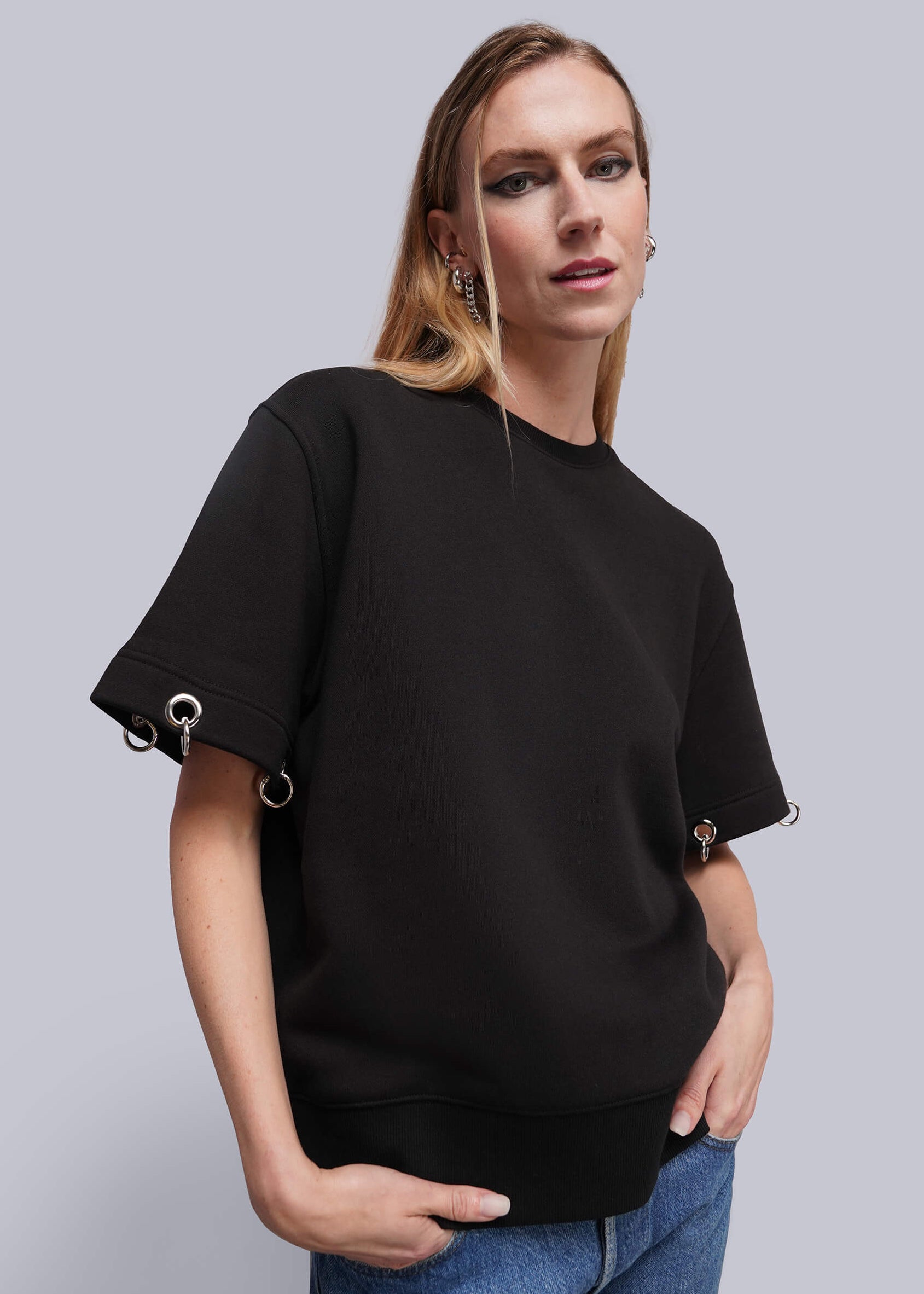 S=A woman modeling a black crewneck sweatshirt with unique sleeve details featuring silver metal rings, paired with blue jeans against a solid gray studio background.