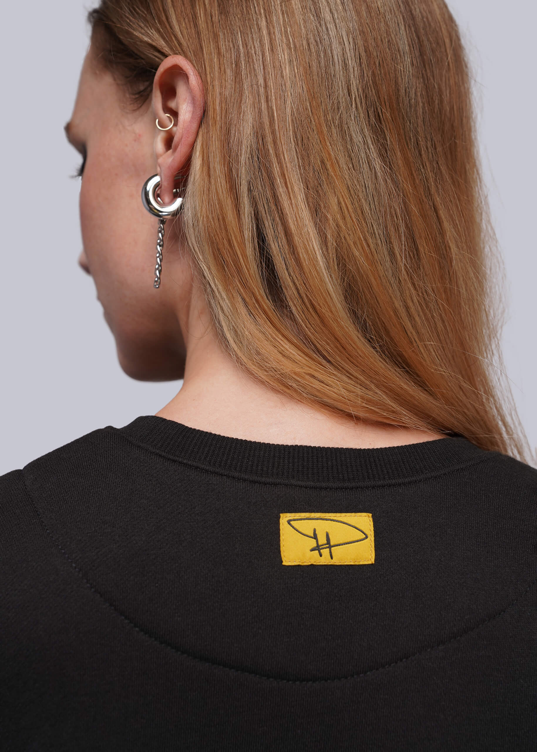 S=Close-up back view of a woman with long blonde hair wearing a black crewneck sweatshirt with a yellow rectangular logo patch on the upper back, and silver hoop earrings.