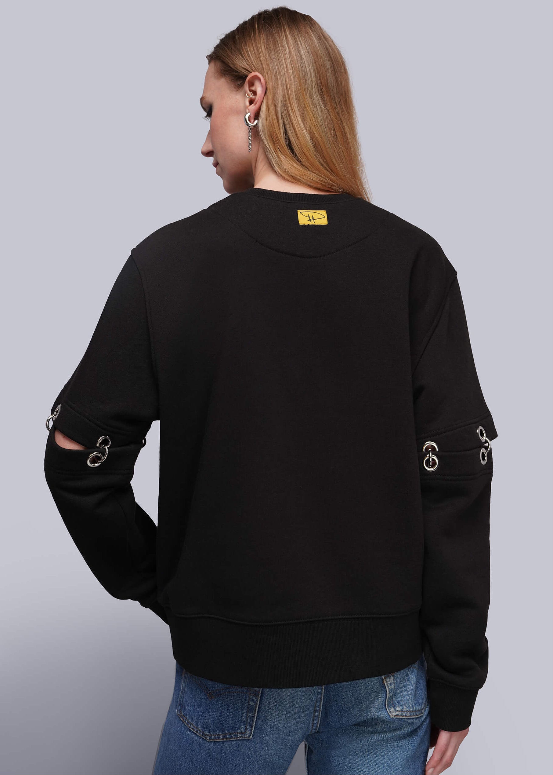 S=A woman shown from the back wearing a black crewneck sweatshirt with silver metal grommet and ring details on the sleeves and a small yellow label at the back neckline, paired with blue jeans against a light gray studio background.