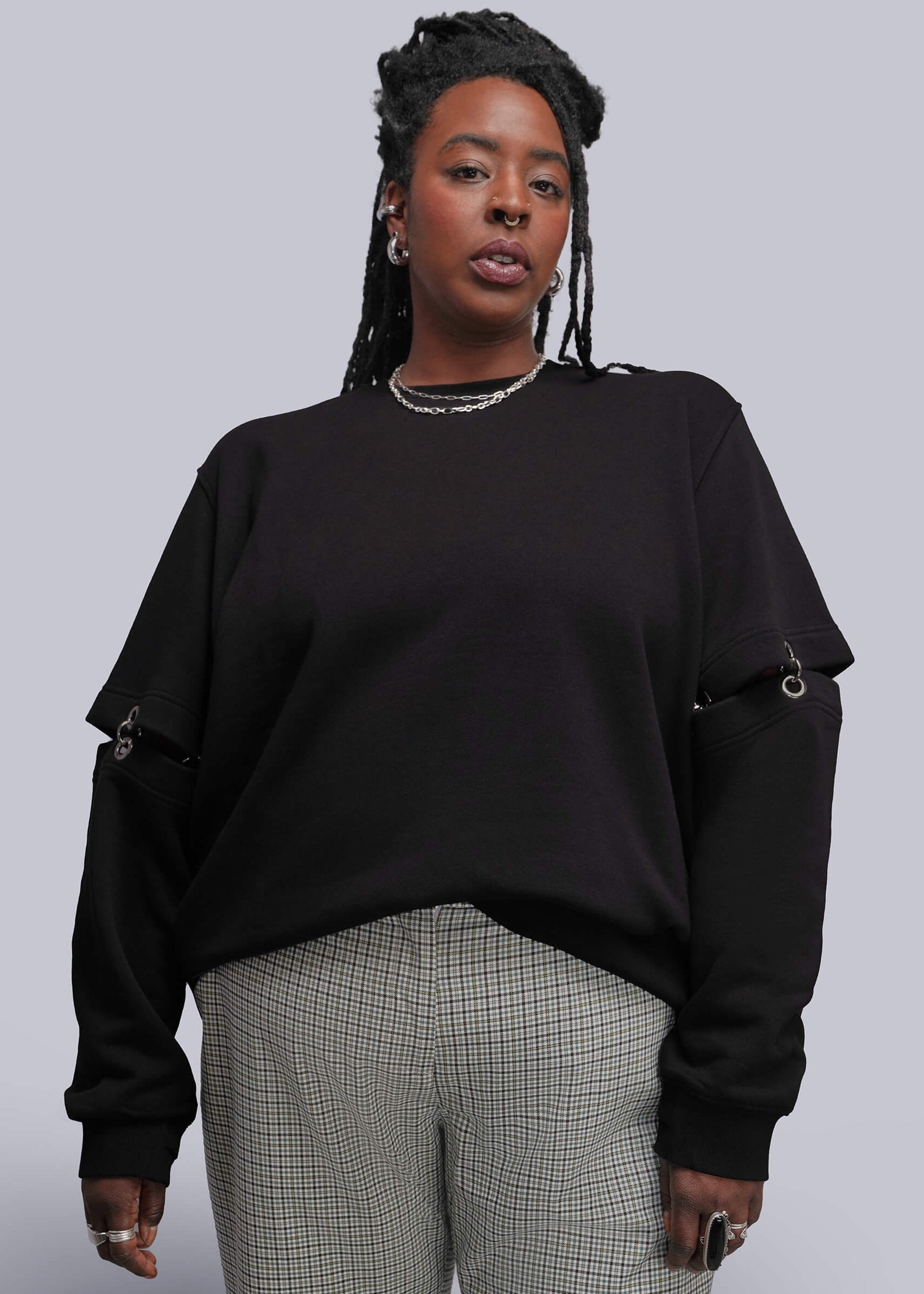 1X=A woman modeling a black crewneck sweatshirt with unique sleeve cutouts and chain details, paired with gray and white checkered trousers, against a solid gray studio background.
