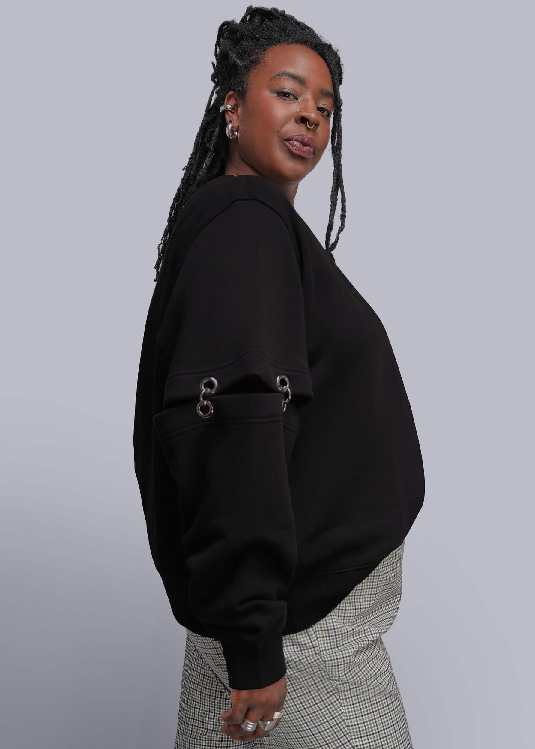 1X=A woman wearing a black cropped sweatshirt with silver grommet details on the sleeves, paired with gray and white checkered pants, photographed against a solid gray studio background.