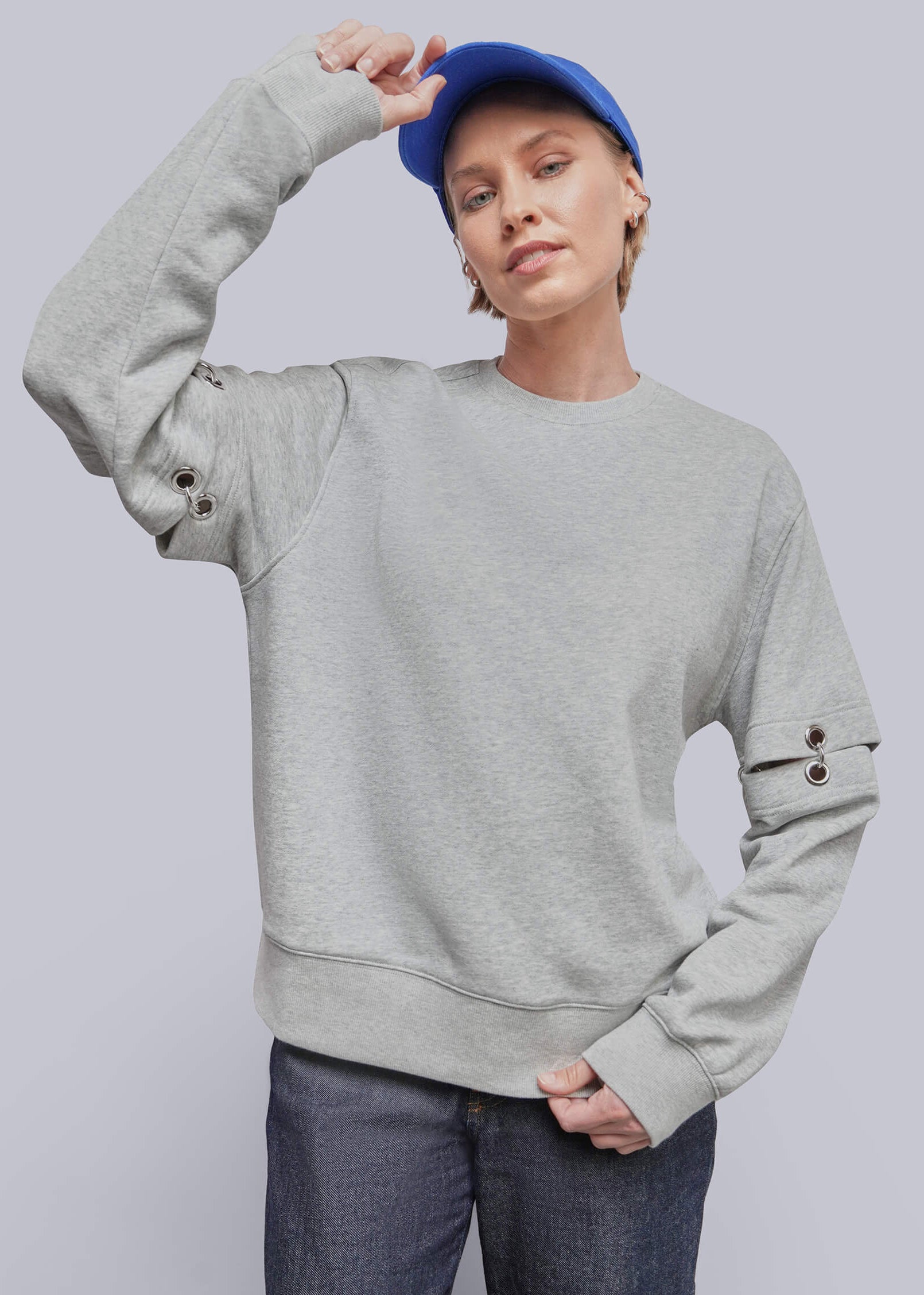 A model wearing a light gray crewneck sweatshirt with decorative button details on the sleeves, paired with dark blue jeans and a blue baseball cap, against a solid gray studio background.
