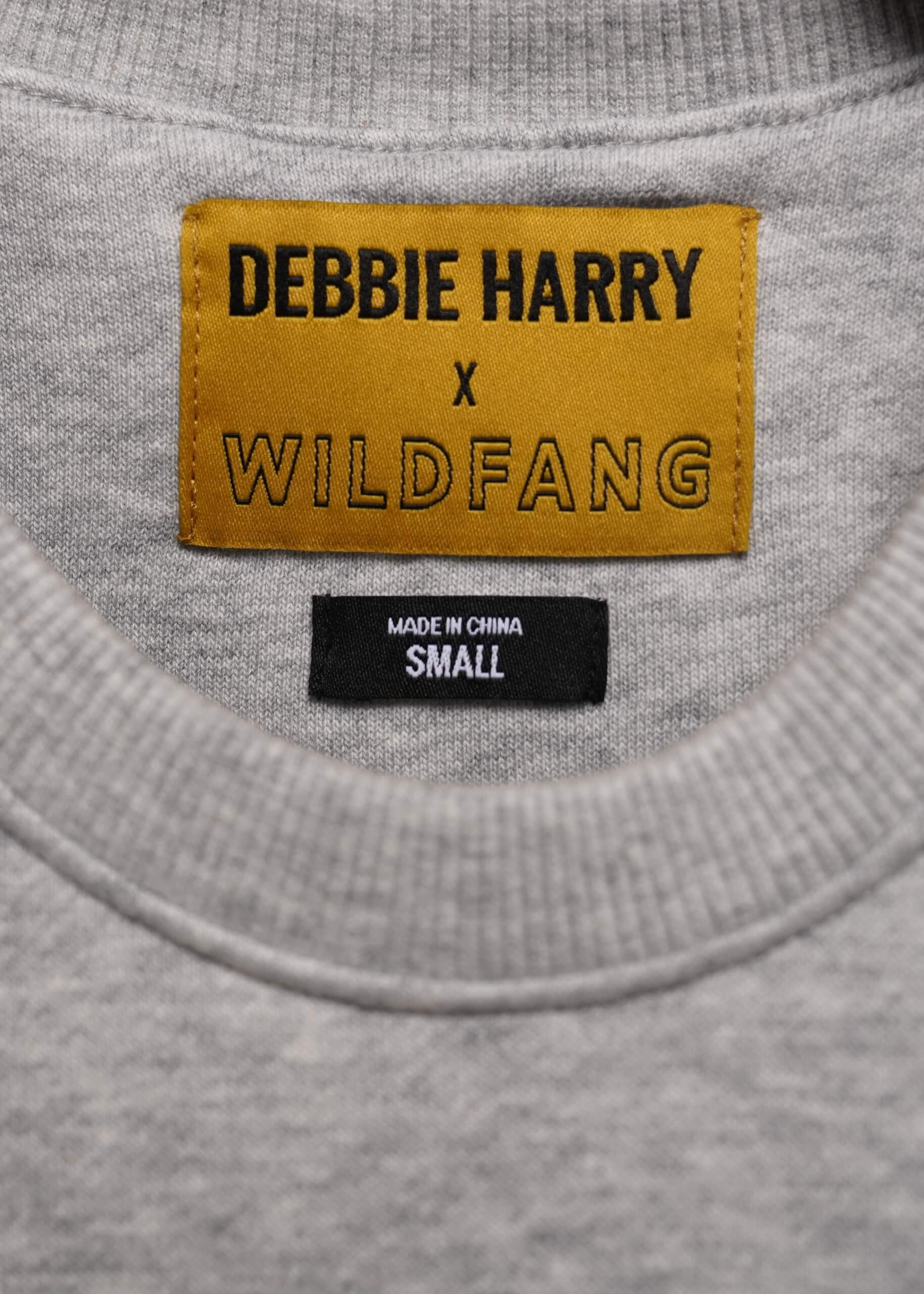 Close-up of a gray heather crewneck sweatshirt showing a yellow woven label that reads 'DEBBIE HARRY x WILDFANG' and a black 'MADE IN CHINA SMALL' tag below it.