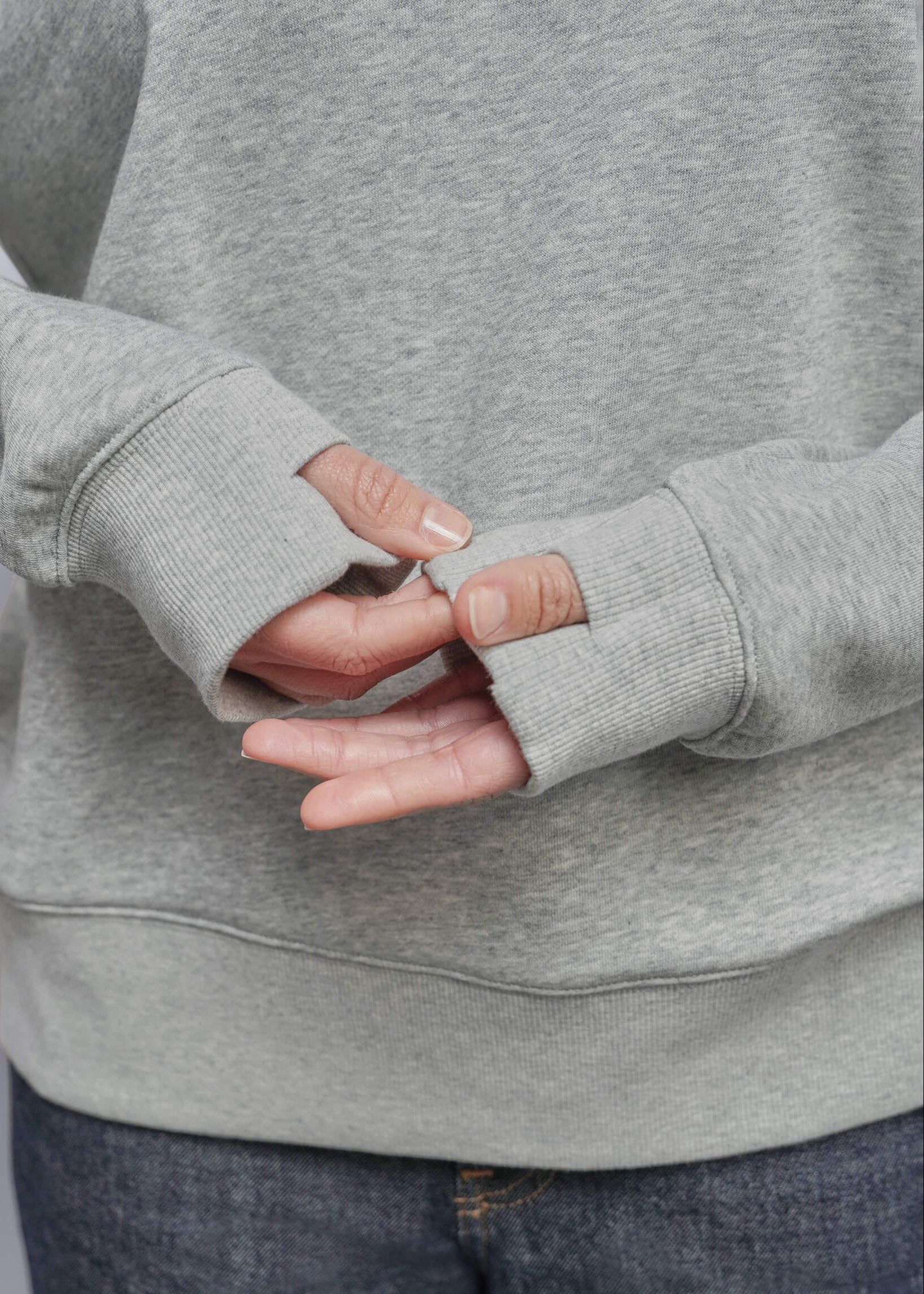 S=Close-up of a person wearing a light gray heather sweatshirt with thumbhole sleeves and dark blue jeans, showing the ribbed cuffs and hands.