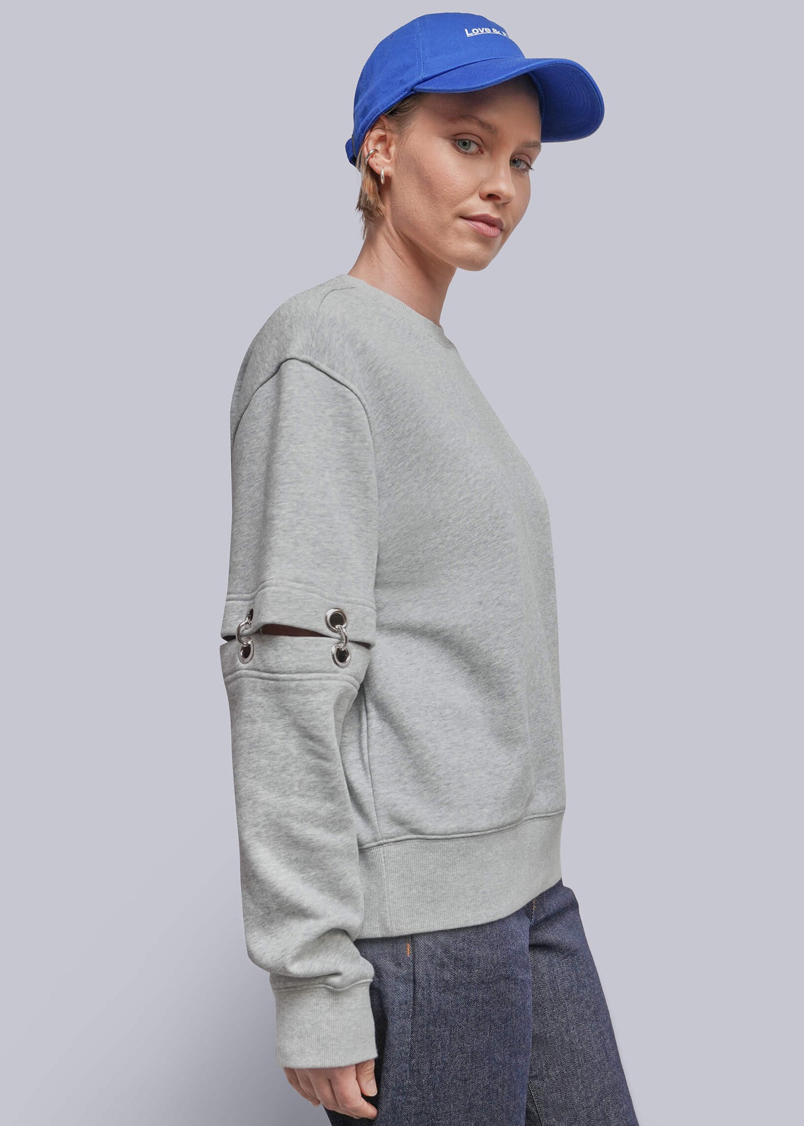 S=A woman in profile wearing a light gray crewneck sweatshirt with unique sleeve details, dark gray wide-leg trousers, and a bright blue baseball cap, set against a solid light gray studio background.