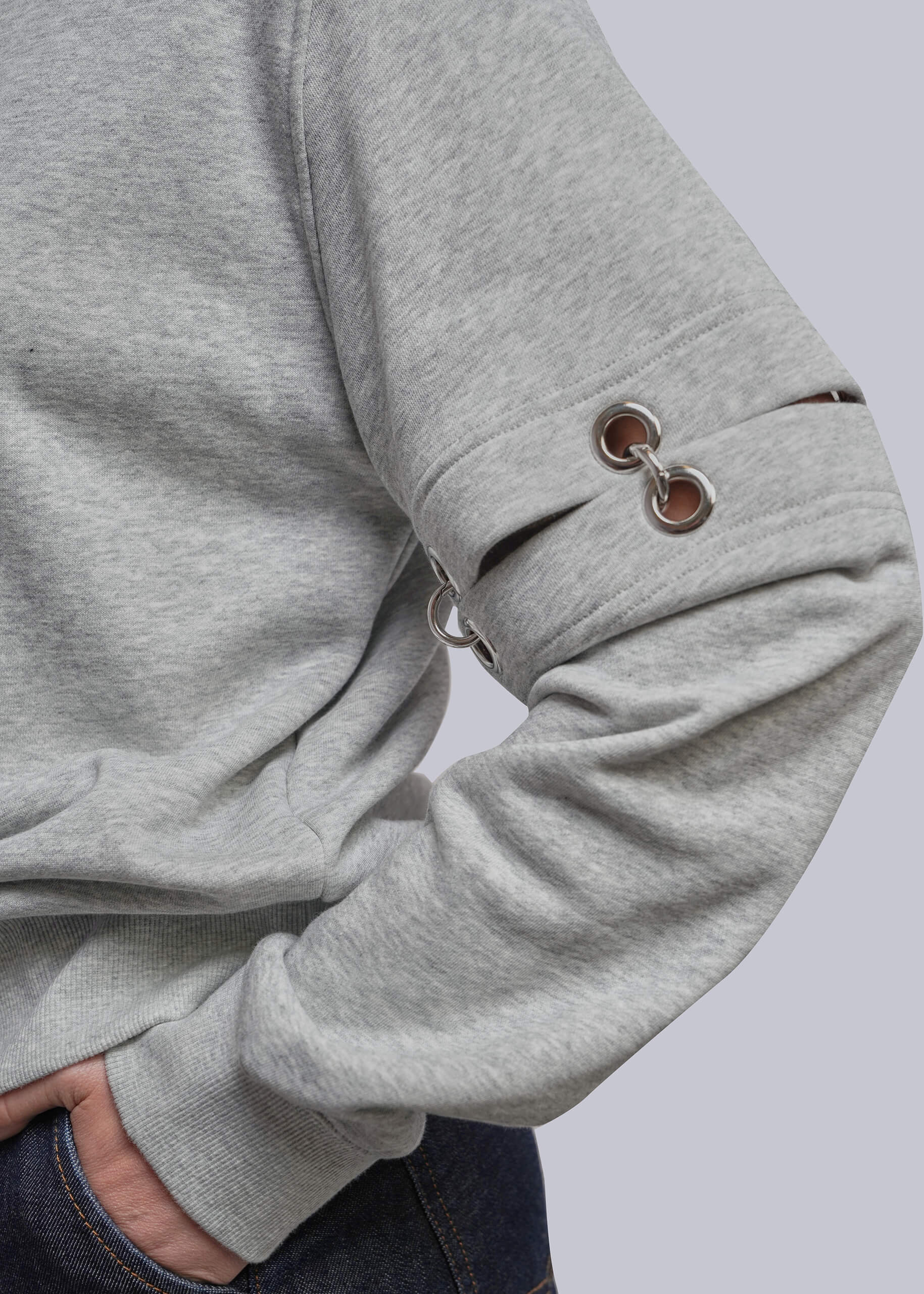 S=Close-up of a person wearing a heather gray long-sleeve sweatshirt with metal grommet details on the sleeve, paired with dark blue jeans against a light gray background.