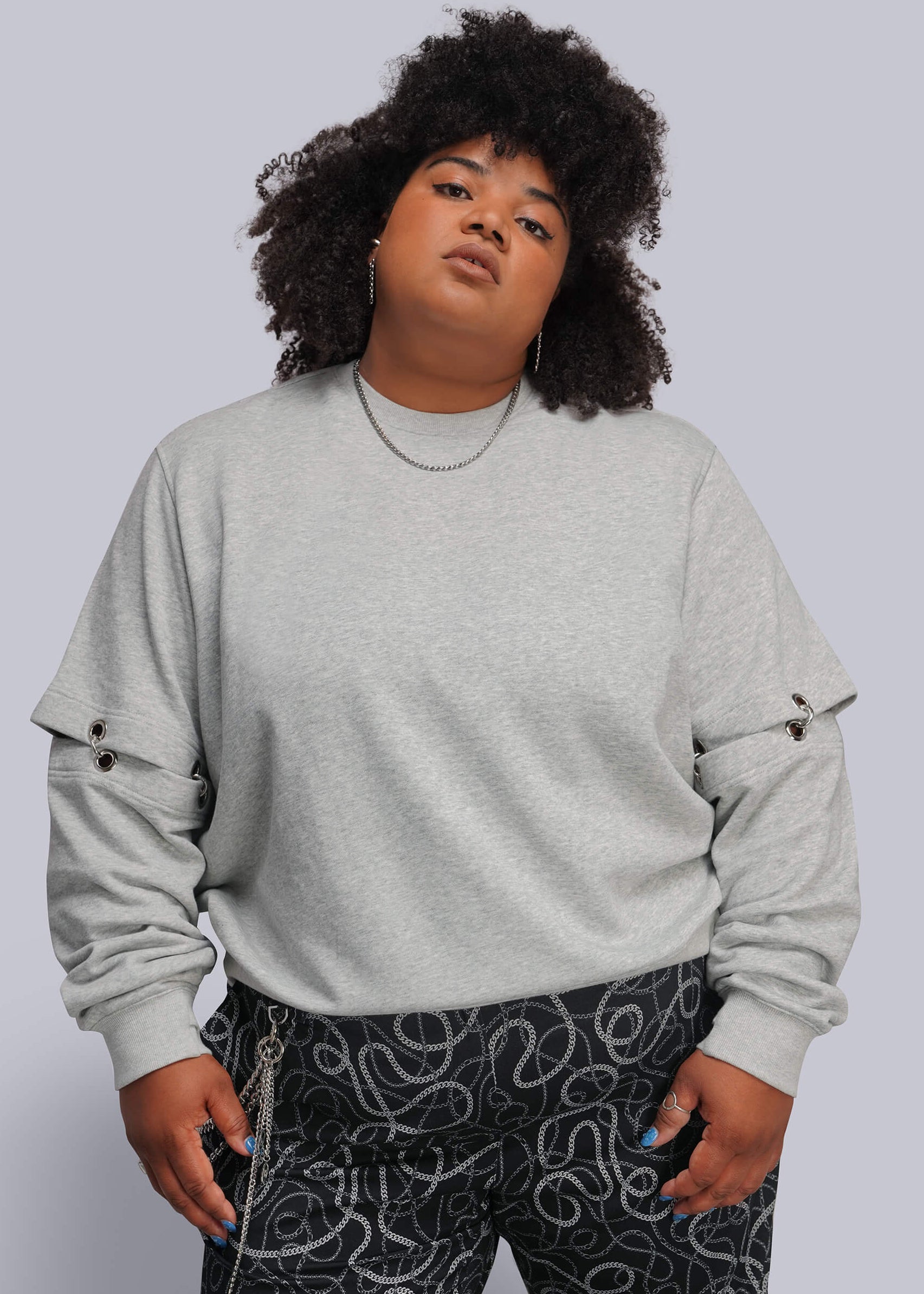 1X=A plus-size model with curly black hair wearing a light gray crewneck sweatshirt with grommet details on the sleeves and black pants with a white abstract pattern, set against a solid light gray background.