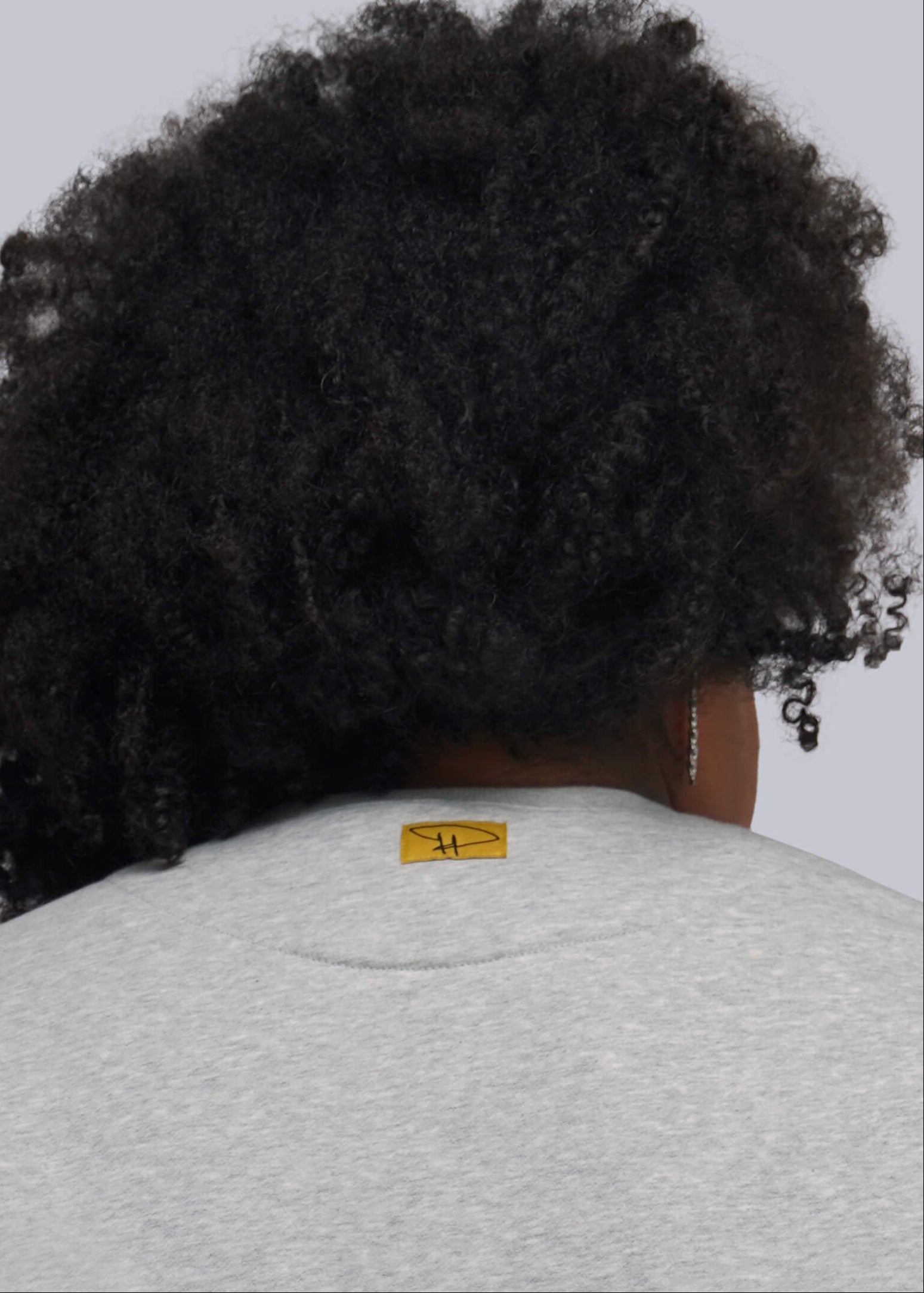1X=Back view of a person with dark curly hair wearing a light gray heather crewneck sweatshirt with a small yellow tag on the outside neck.