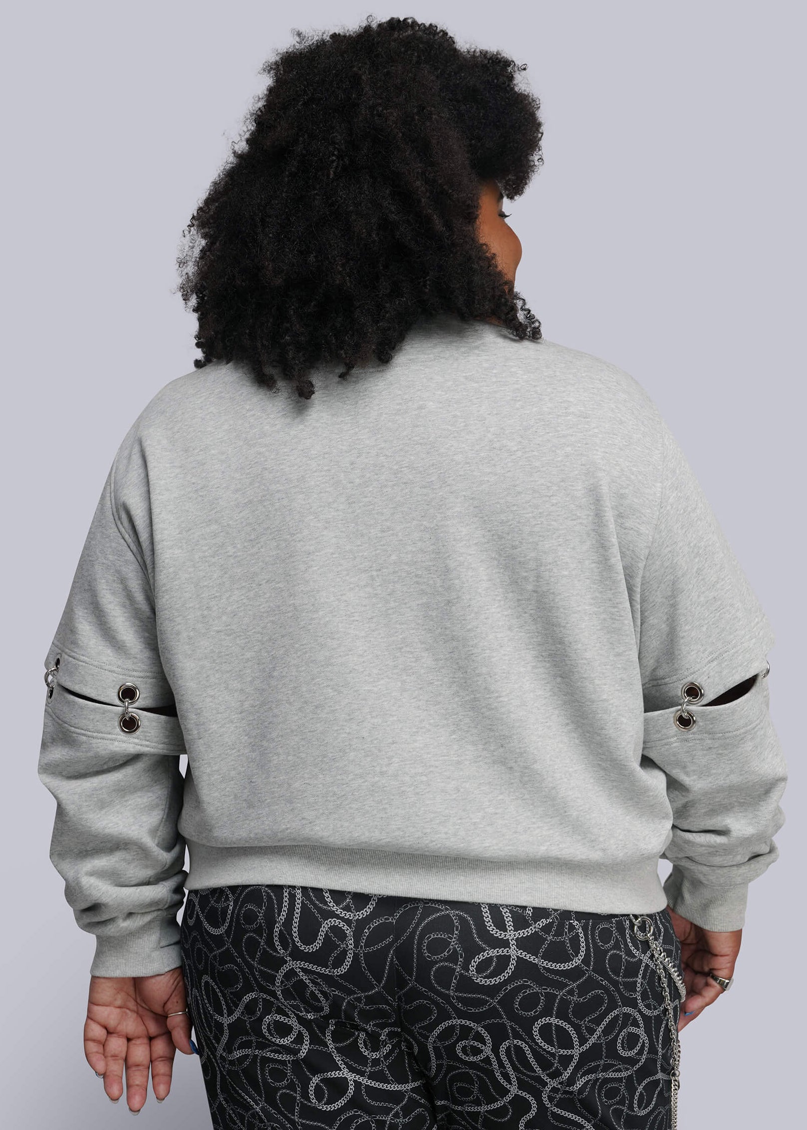 1X=A person shown from the back wearing a light gray heather sweatshirt with decorative black button details on the sleeves and dark patterned pants, against a solid light gray background.