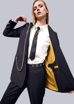 S=A stylish person poses confidently in a black suit with a white shirt and tie, showcasing the Debbie Harry X Wildfang Double Breasted Blazers striking yellow lining. The outfit is perfectly accessorized with chains and exquisite metal detailing.