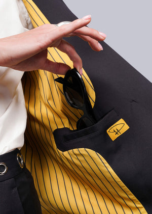 S=Black sunglasses are slipped into the inner pocket of a Debbie Harry X Wildfang Double Breasted Blazer, with yellow and black striped lining featuring an eyeglass frame logo. A person pairs it with a white shirt and dark pants adorned with metal detailing.