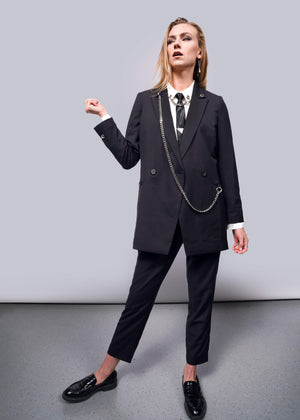 S= Confidently posing against a plain backdrop, she embodies elegance in a black tailored suit from the Wildfang collection. Her white shirt complements the Debbie Harry X Wildfang Double Breasted Blazers chain accessory and metal detailing, while black loafers complete her bold, sophisticated look.