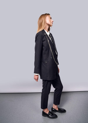 S=A person with long hair in profile against a plain background wears a Debbie Harry X Wildfang Double Breasted Blazer with metal detailing. They pair it with shiny black loafers, looking upward, their hands relaxed at their sides.