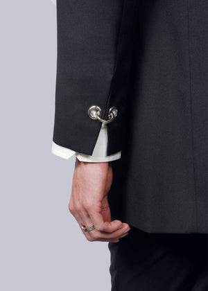 S= A person wears the sleek black Debbie Harry X Wildfang Double Breasted Blazer featuring metal hoop cufflinks while holding the lapel. A white shirt sleeve shows, paired with a silver ring on their middle finger, contrasting against a white background and highlighting striking metal details.